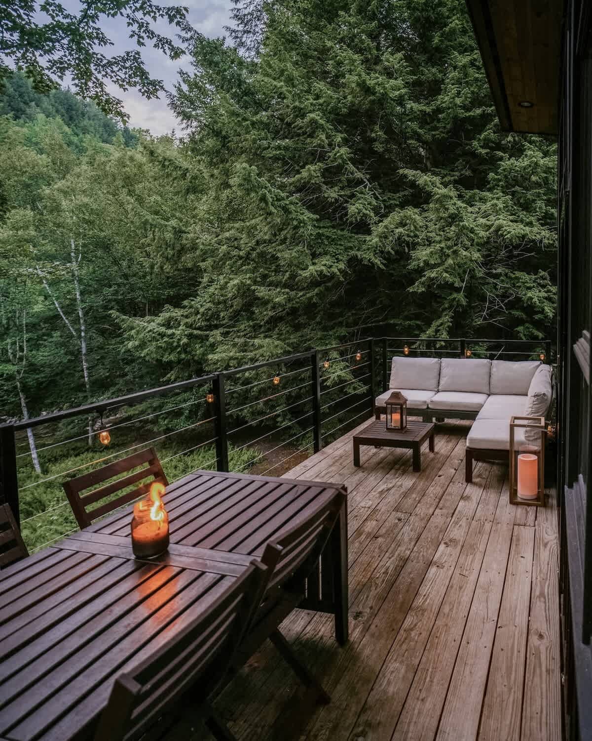 A spacious wooden deck overlooks a lush green forest, featuring a dark wood dining table with chairs and a light-colored sectional sofa with a coffee table. String lights and lanterns add a warm ambiance to the outdoor seating area.
