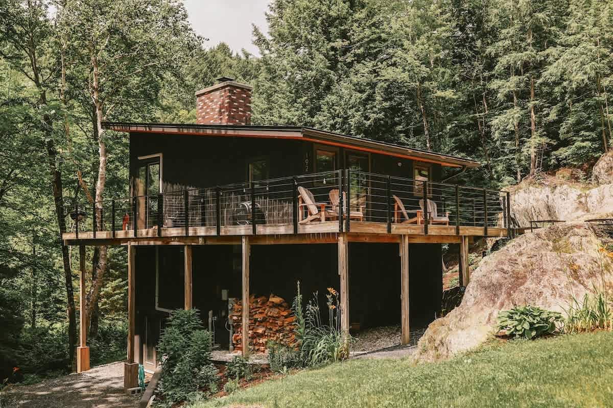 The dark-sided house from another angle, showcasing a multi-level wooden deck, a brick chimney, and a neatly stacked pile of firewood underneath, surrounded by lush greenery.