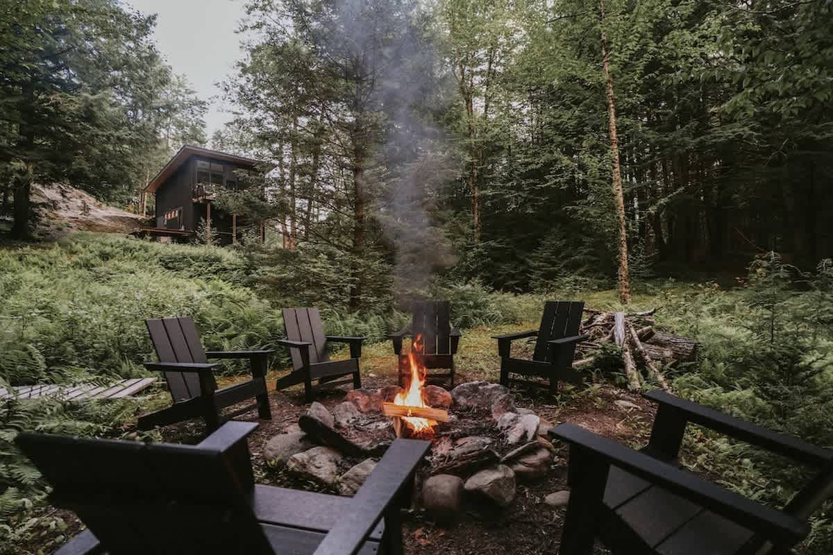 An inviting outdoor fire pit area with a roaring fire, surrounded by dark Adirondack chairs, set in a clearing within a lush forest with the house visible in the background.