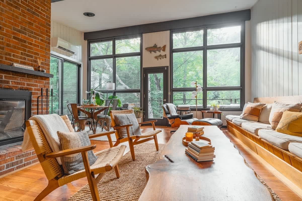 Spacious living room with a brick fireplace, large floor-to-ceiling windows offering forest views, and a mix of seating including wooden armchairs, a long built-in sofa, and a live-edge coffee table.
