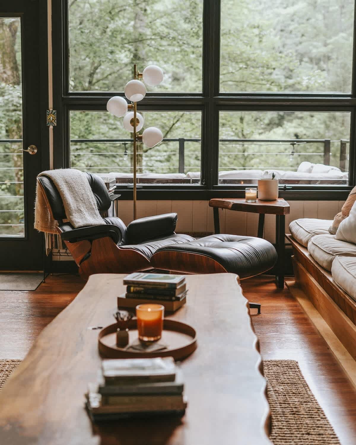 A comfortable black leather lounge chair with a matching ottoman is positioned by large windows offering serene forest views. A modern floor lamp provides ambient lighting, and a rustic live-edge coffee table is in the foreground.