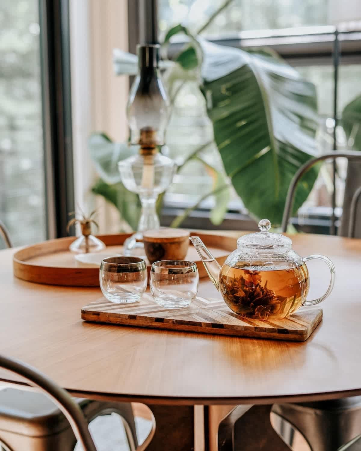 A round wooden dining table set with a glass teapot containing blooming tea, two glass cups, and a wooden tray. Large windows and lush greenery are visible in the background.