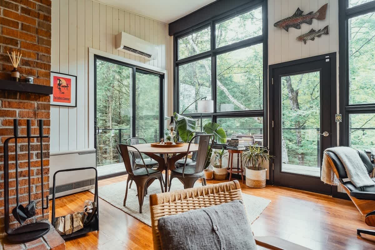A bright dining area features a round wooden table with four industrial-style metal chairs, set on a light rug. Large floor-to-ceiling windows offer expansive views of lush green trees, while a brick fireplace adds a rustic touch to the left.
