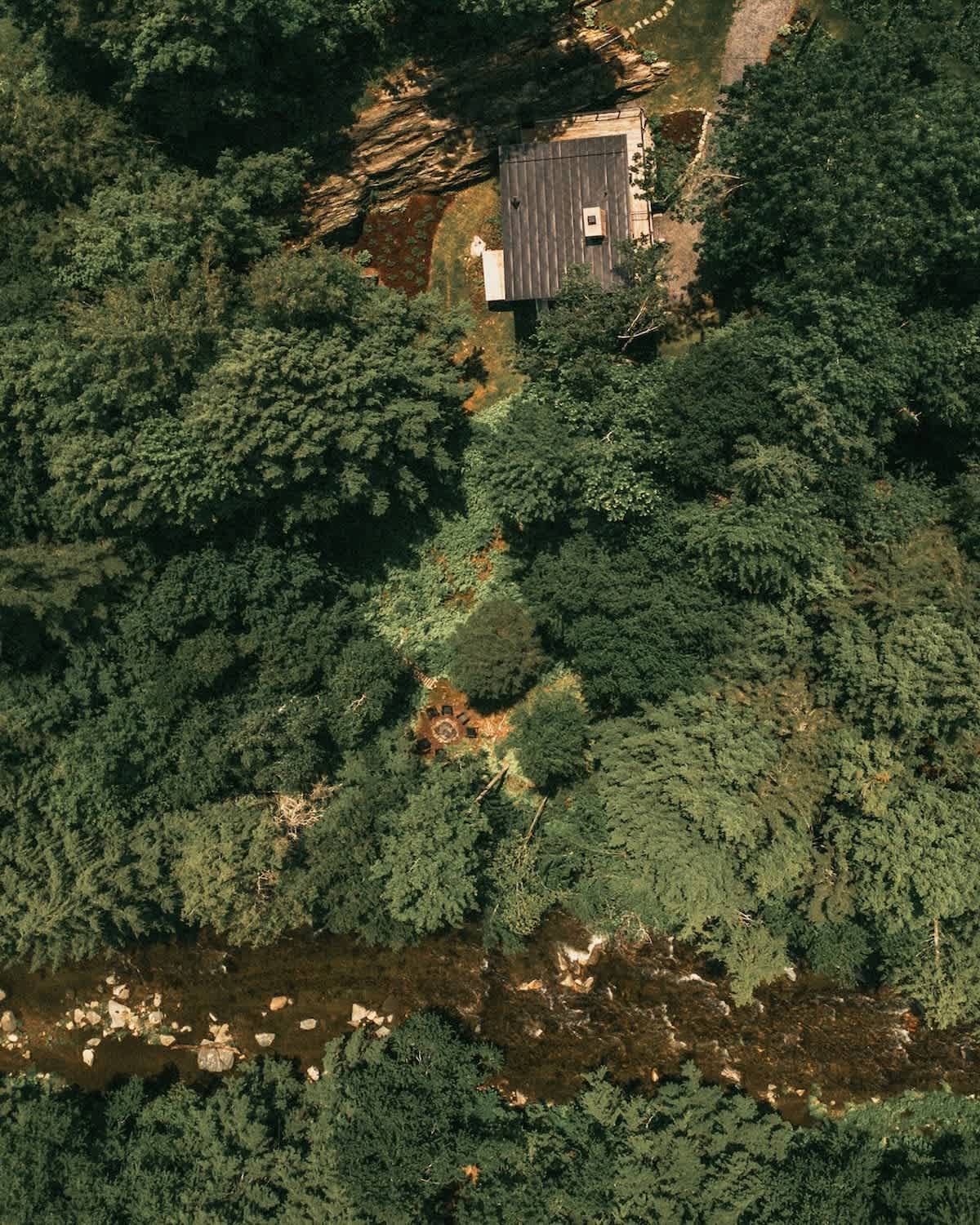 An aerial view shows a dark-roofed cabin surrounded by dense green forest, with a winding river visible at the bottom left. A small clearing with a fire pit is nestled among the trees near the cabin.
