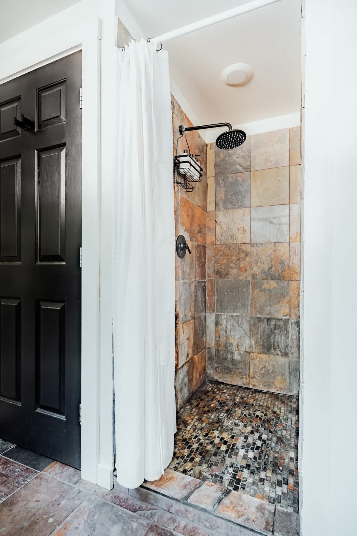 A shower stall with multi-toned slate tile walls and a mosaic tile floor, featuring a black rain shower head and a white shower curtain.