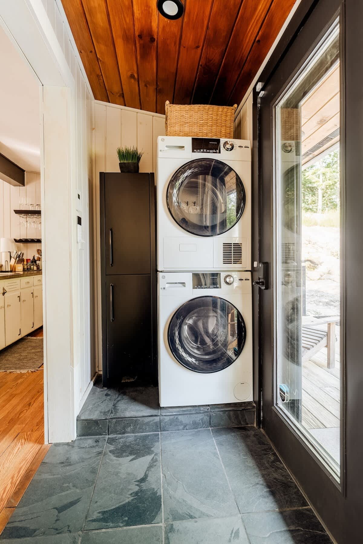 A dedicated laundry area with a stacked white washer and dryer unit, a tall black cabinet, and a glass-paned door leading to the exterior. The ceiling is paneled with warm wood.