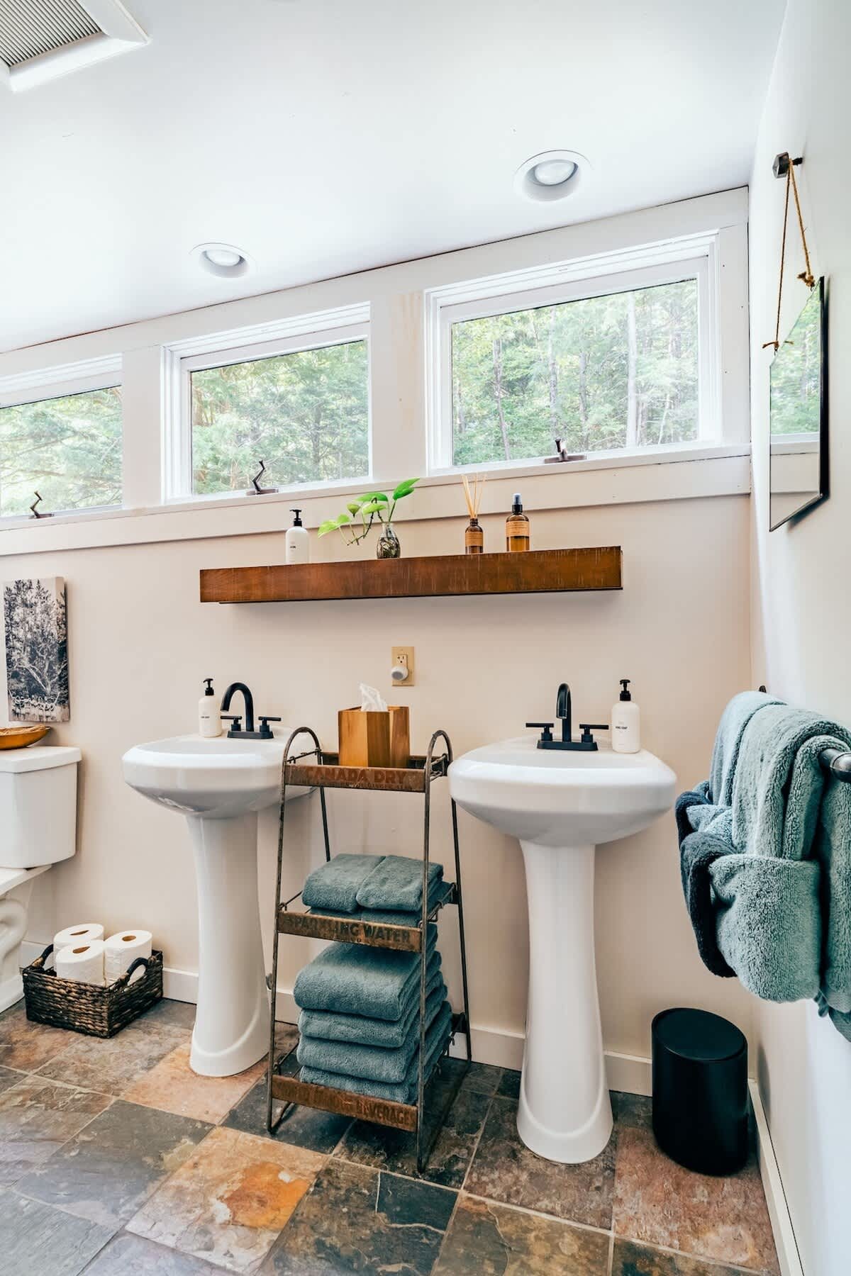 A spacious bathroom with two white pedestal sinks, each with a black faucet, a wooden shelf above, and multiple windows offering natural light. The floor is tiled with multi-colored slate.