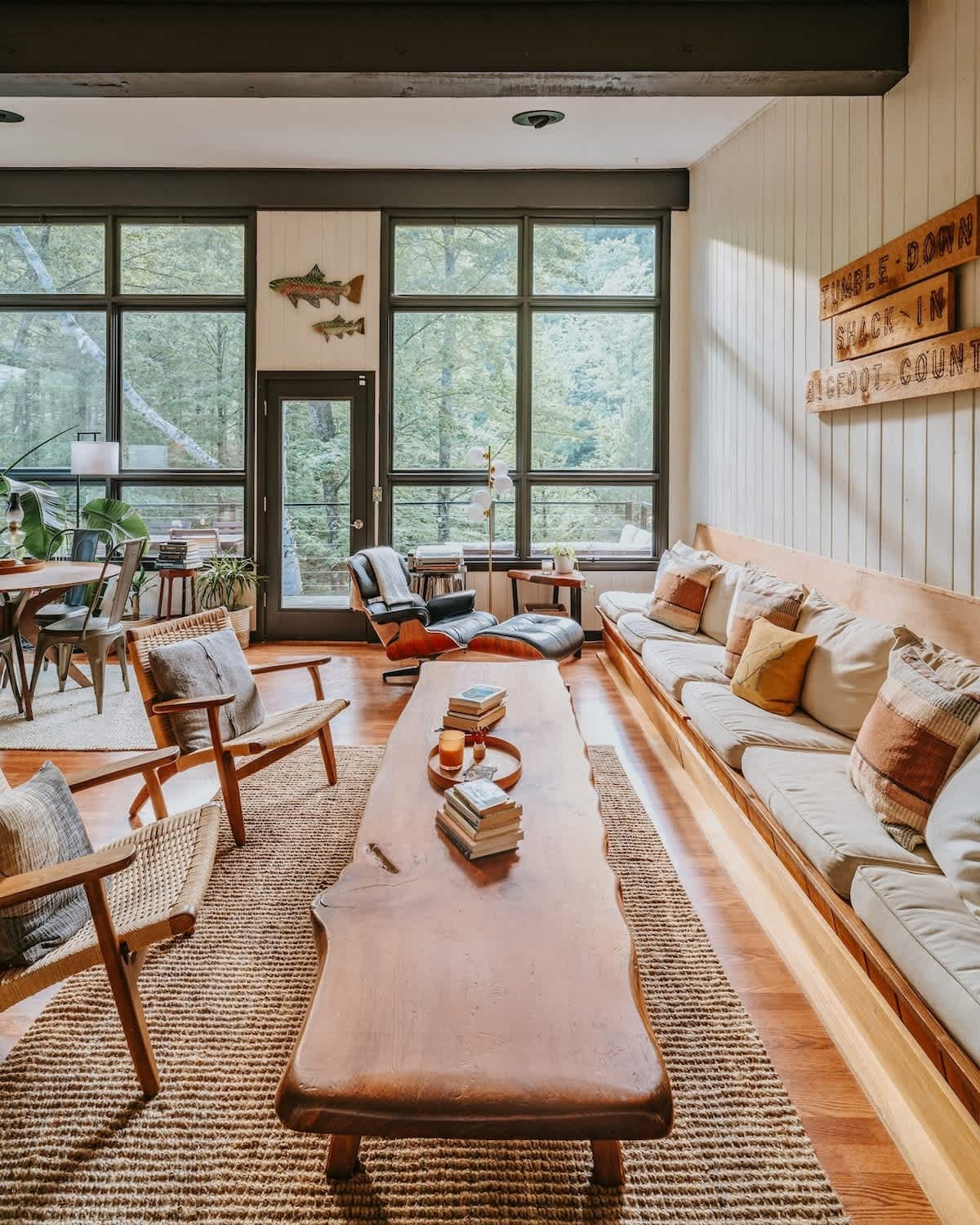 Expansive living and dining area bathed in natural light from large floor-to-ceiling windows overlooking a forest. Features a long built-in sofa, a rustic live-edge coffee table, and a black leather lounge chair, all on warm hardwood floors with a jute rug.