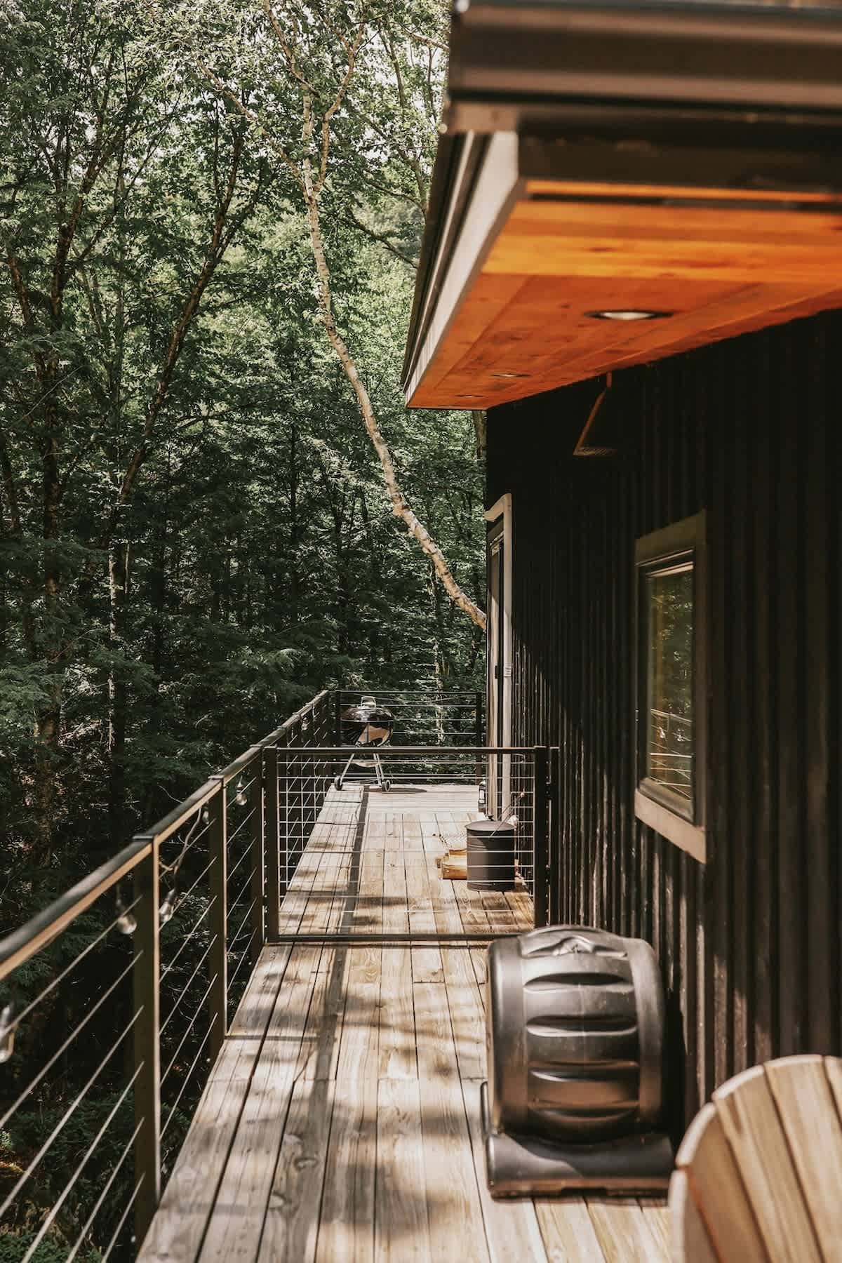 A narrow wooden deck runs along the side of the black cabin, offering views of the surrounding dense forest. A barbecue grill and an outdoor fan are visible on the deck, which is enclosed by black metal railings.
