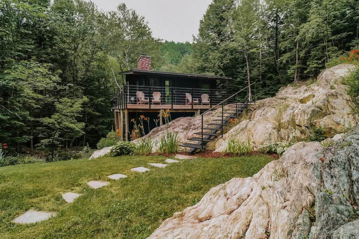 A modern black cabin with a raised wooden deck is set against a backdrop of dense green forest and large rock formations. A stone path and metal staircase lead up to the property, surrounded by grass and wildflowers.