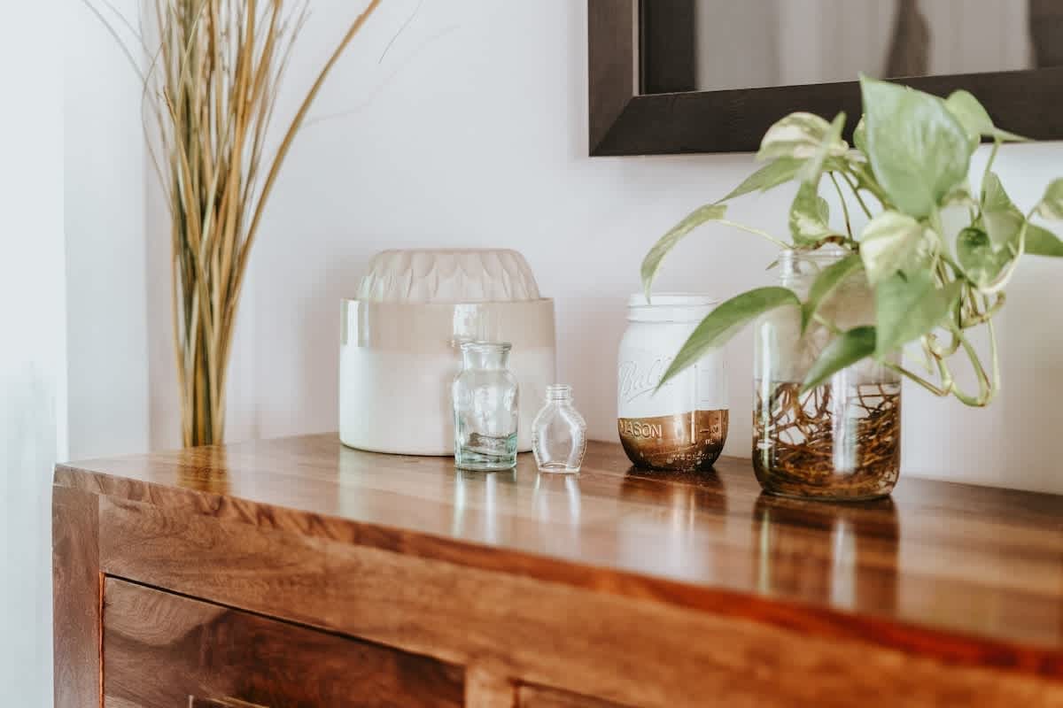 A close-up of a polished wooden console table displaying various decorative items, including dried grasses in a vase, small glass bottles, and a mason jar with a thriving green plant, beneath a framed mirror.