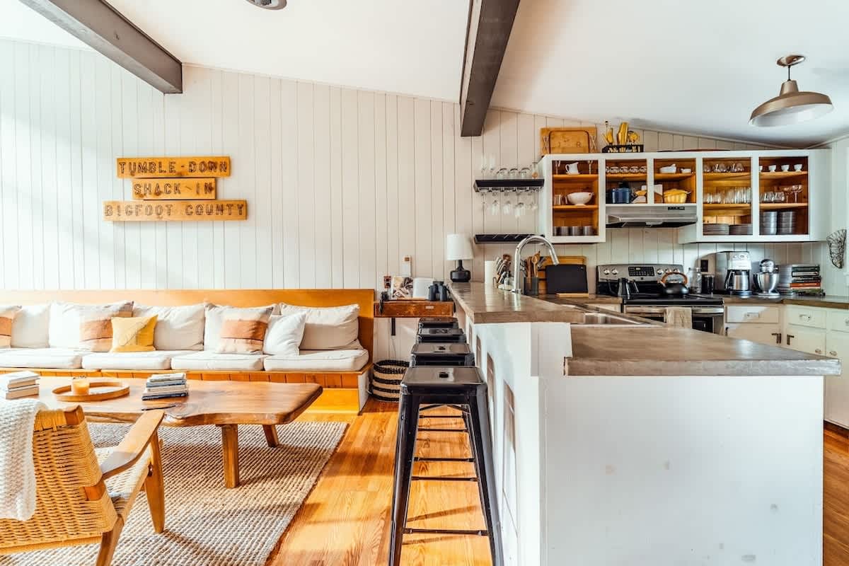 Bright kitchen area with white vertical paneling, a concrete-topped island with seating, and white open-shelf cabinets, flowing into the living space with a built-in sofa and a large wooden coffee table.