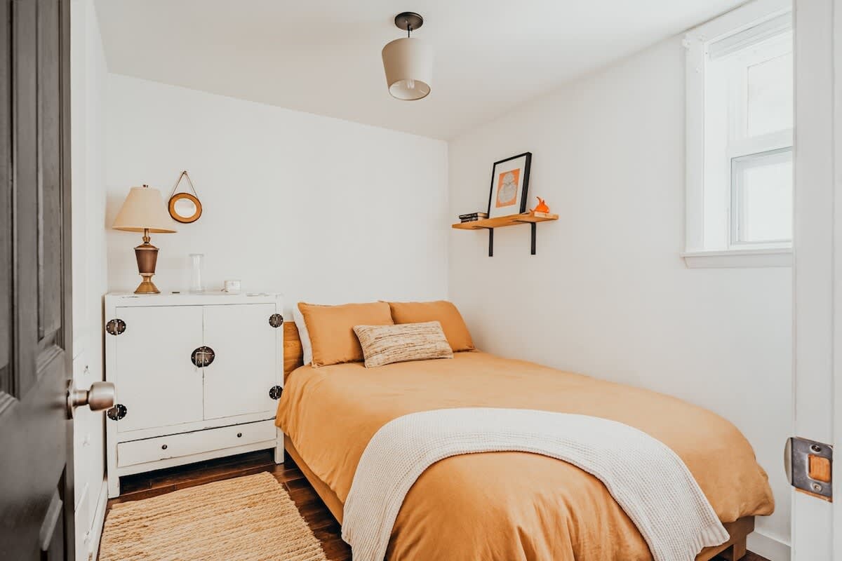 A cozy bedroom with white walls, a twin bed featuring mustard yellow bedding and a white throw, a white dresser with dark hardware, and a small window providing natural light. A light fixture hangs from the ceiling.