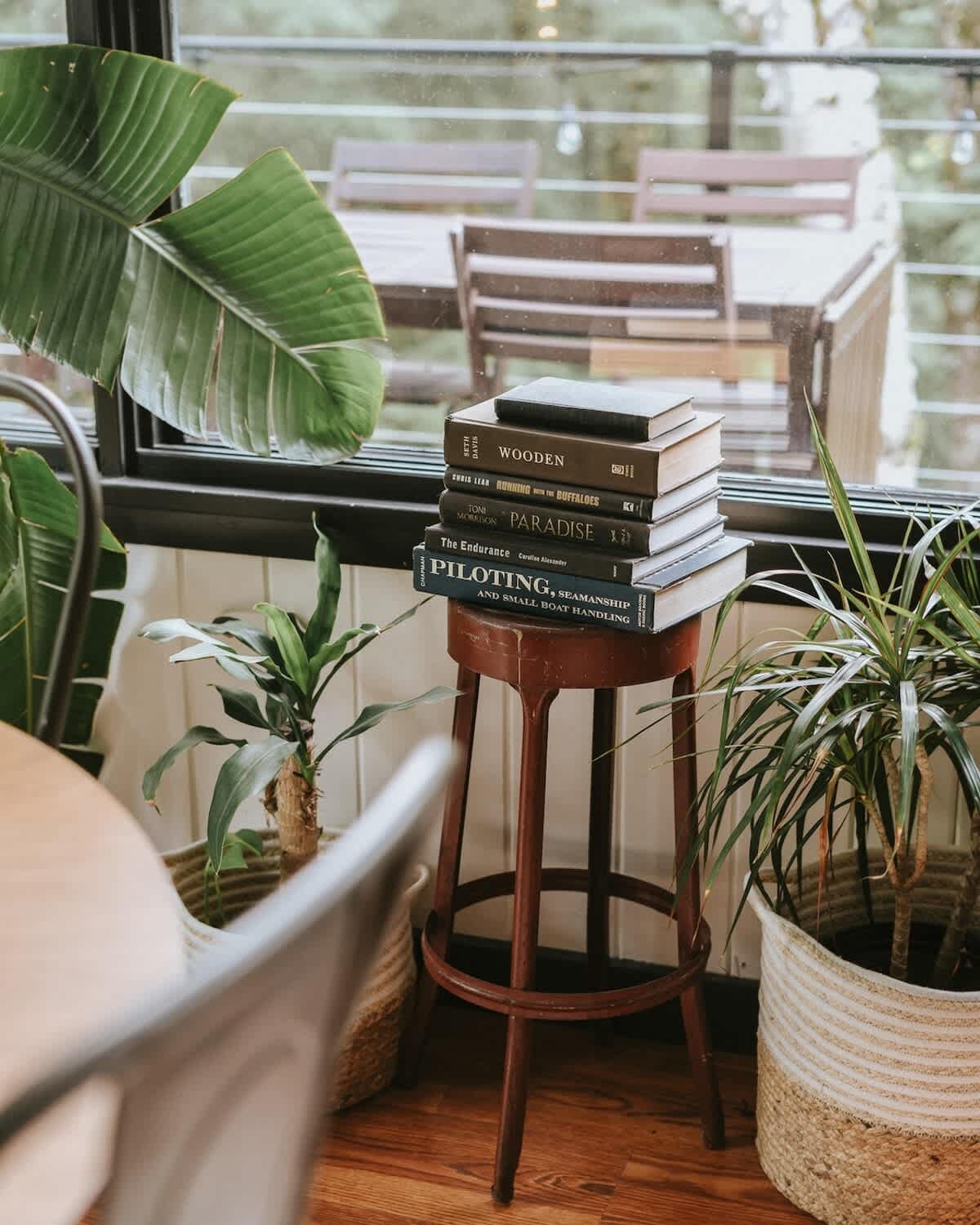 A close-up of a rustic wooden stool holding a stack of books and surrounded by potted plants, positioned by a large window offering glimpses of the outdoor scenery.