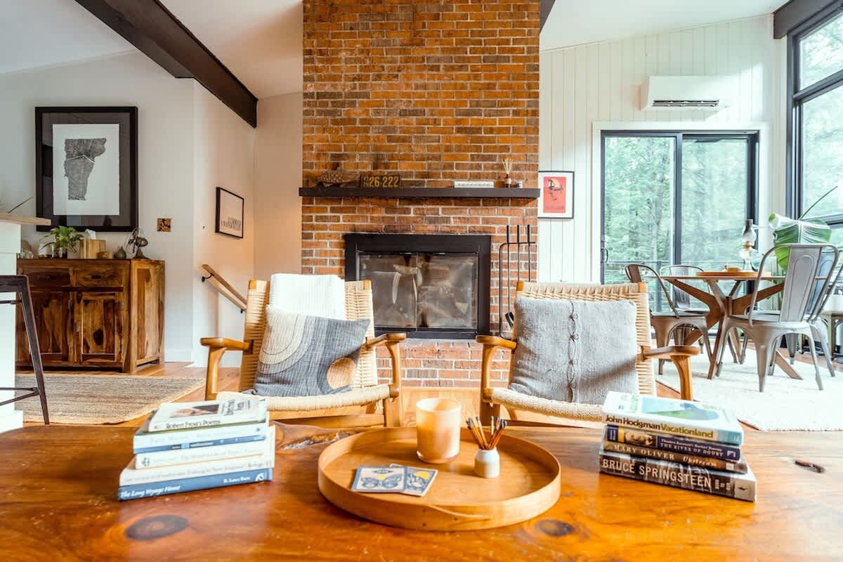 Warm living room centered around a brick fireplace with a wooden mantel, flanked by two woven wooden armchairs with throw pillows, and a large live-edge coffee table adorned with books and a tray.