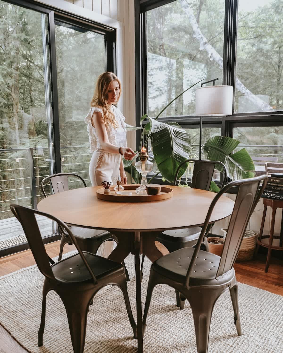 A woman lights a candle on a wooden tray centerpiece on the round dining table, surrounded by industrial-style metal chairs. The area is brightly lit by expansive floor-to-ceiling windows that reveal a lush outdoor forest.