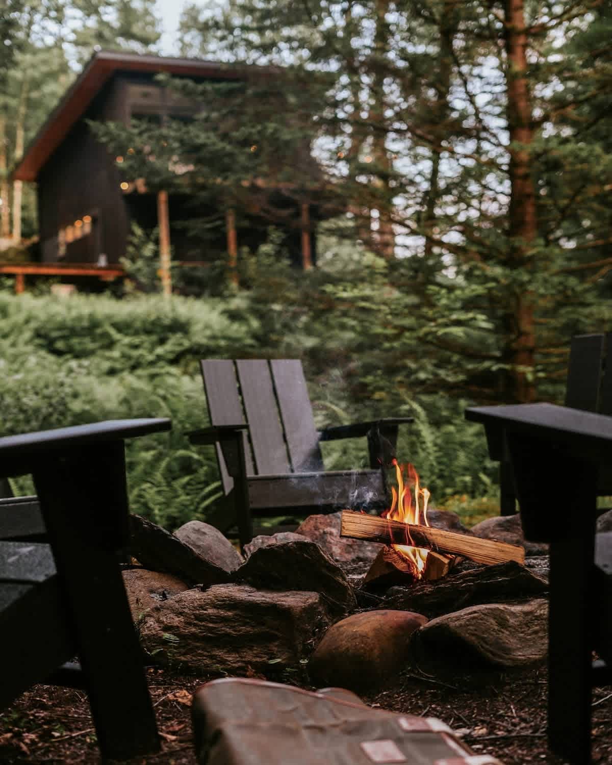 A close-up view of a crackling fire in a stone fire pit, with dark Adirondack chairs arranged around it and the house visible in the distance through the trees.