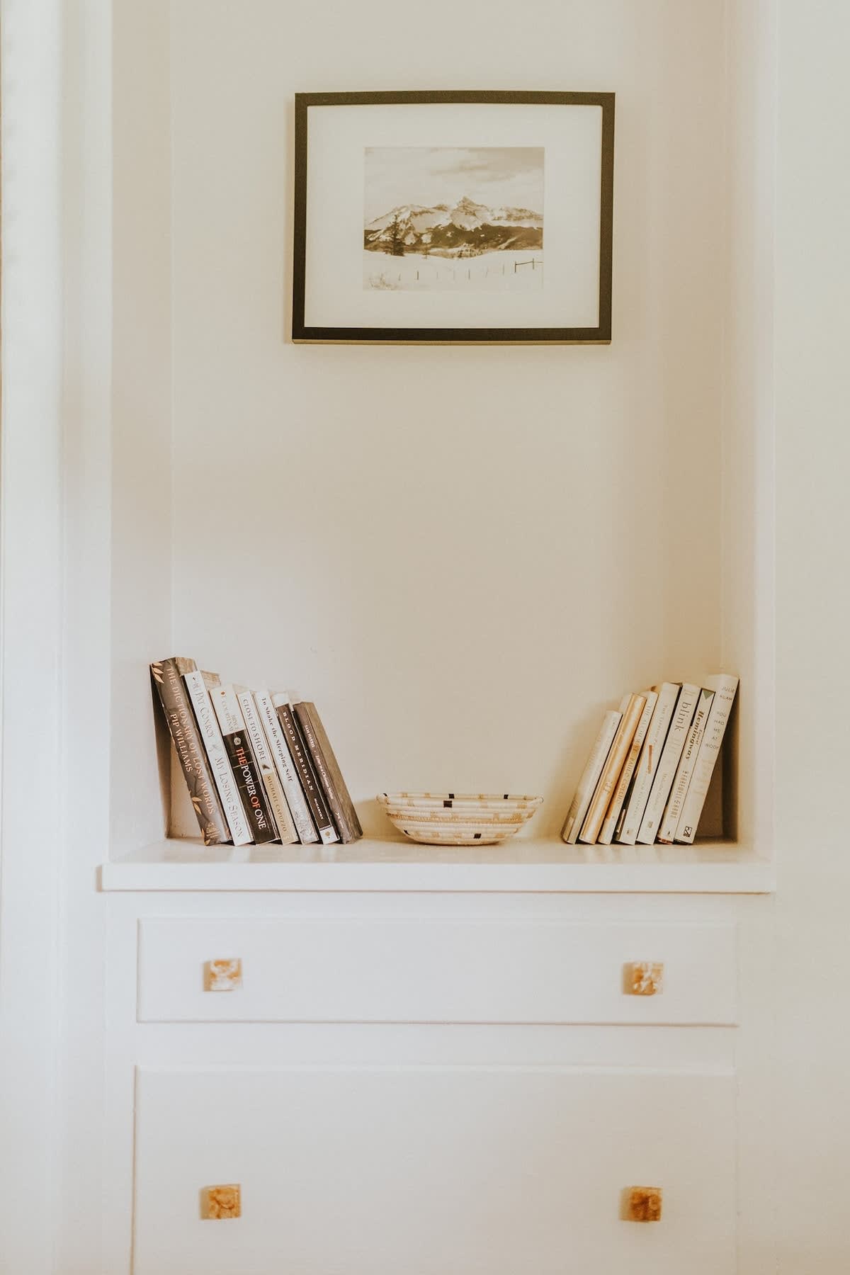 A cozy built-in shelving unit features neatly arranged books and a decorative woven bowl, with two drawers below. A framed black and white landscape print hangs above the alcove, set against a light-colored wall.