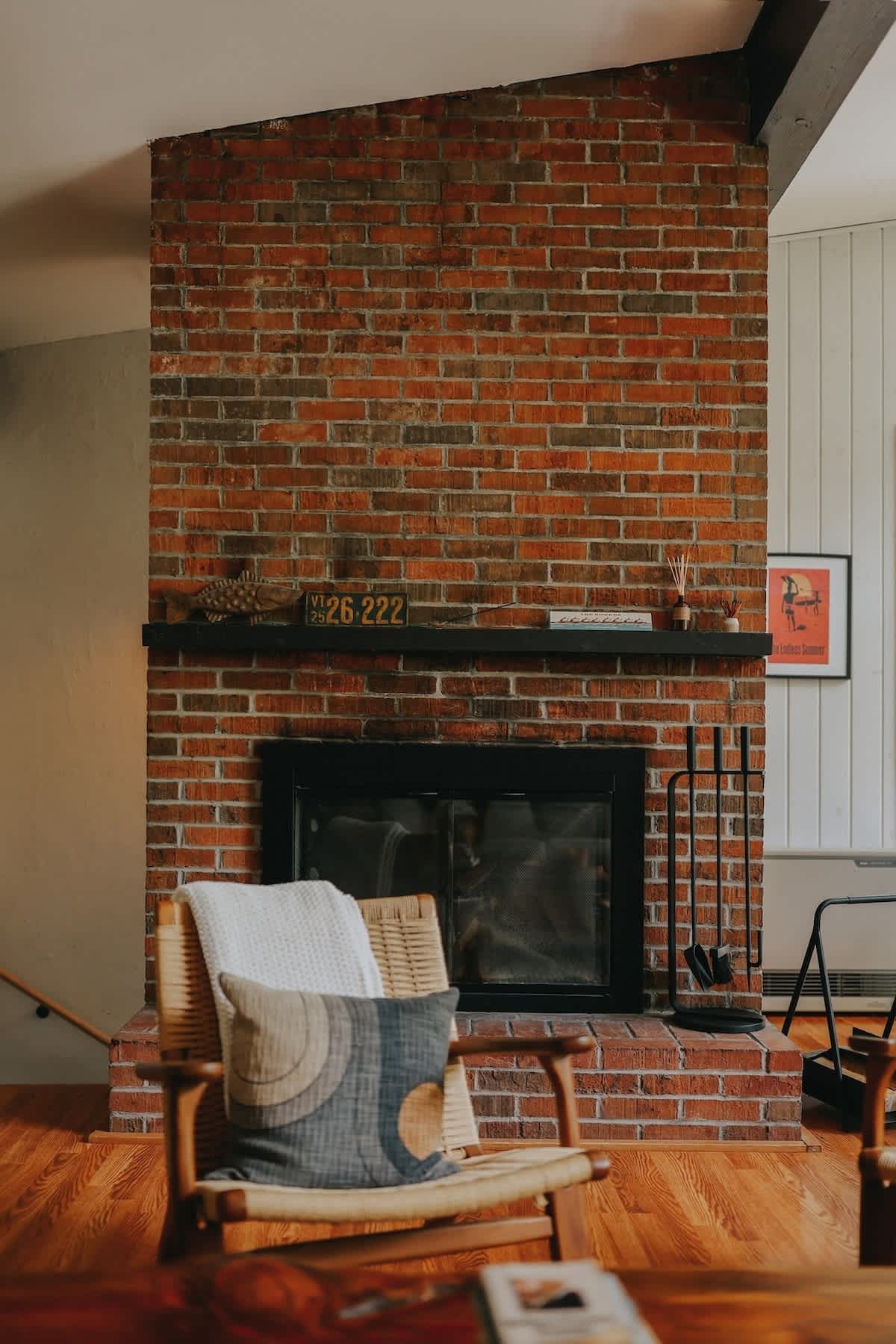 A prominent, floor-to-ceiling brick fireplace with a black mantel and hearth, equipped with fireplace tools. A cozy woven armchair with a patterned pillow sits in the foreground, complementing the warm hardwood floors.