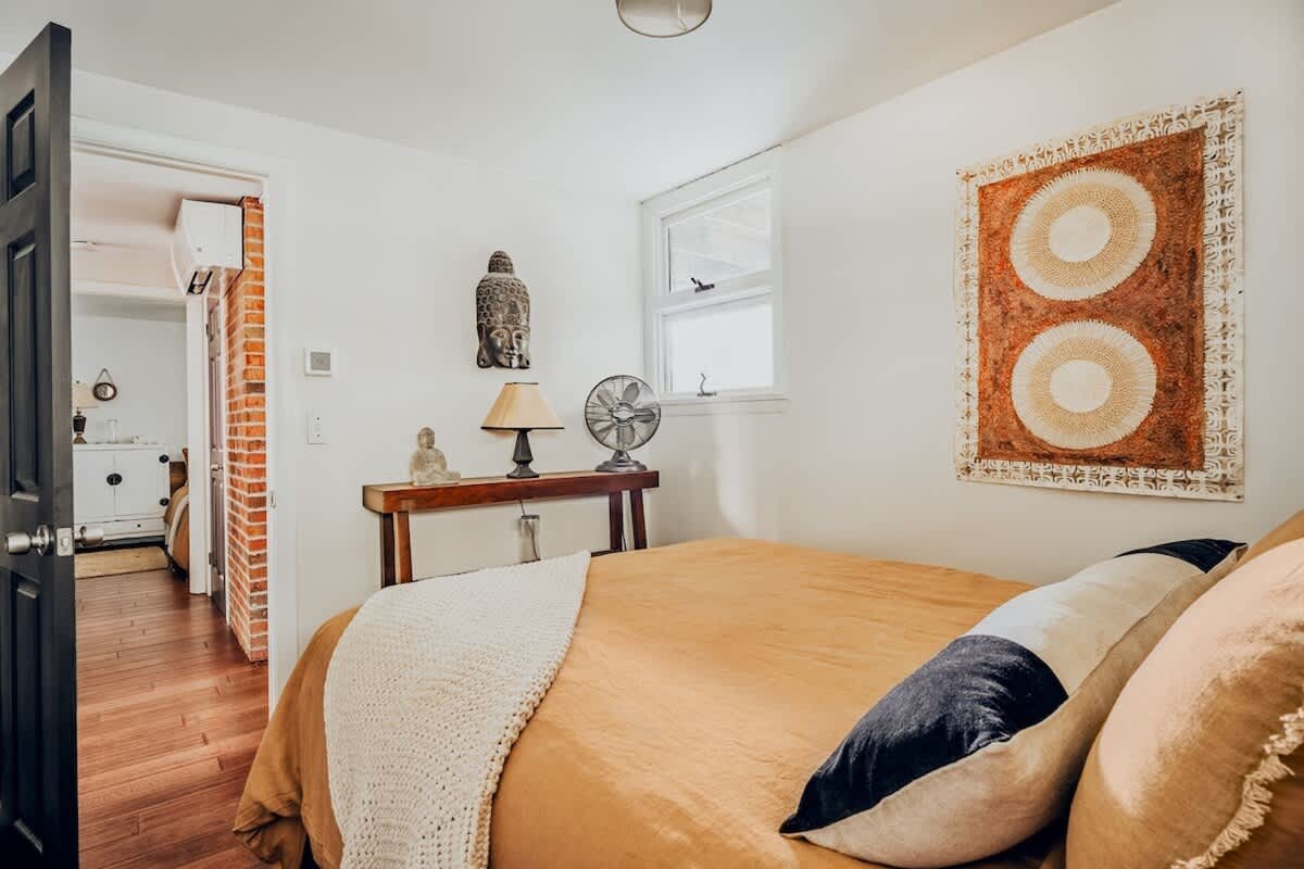 Cozy bedroom featuring a queen-sized bed with mustard yellow bedding and decorative pillows, a small window, and a wooden console table adorned with a Buddha statue and a table lamp.