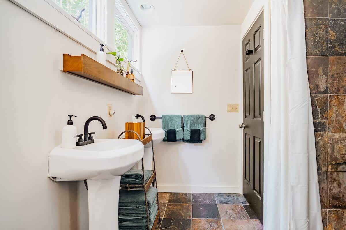 A bright bathroom featuring a white pedestal sink with a black faucet, a wooden shelf above, and a mirror. The floor is tiled with multi-colored slate, and teal towels are neatly stacked and hung.