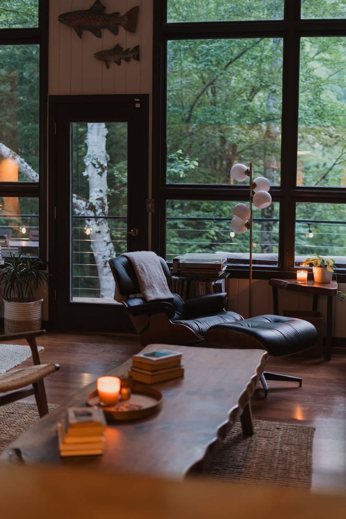 A cozy living room with large floor-to-ceiling windows overlooking a lush forest, featuring a black leather lounge chair with an ottoman, a wooden coffee table, and warm ambient lighting.