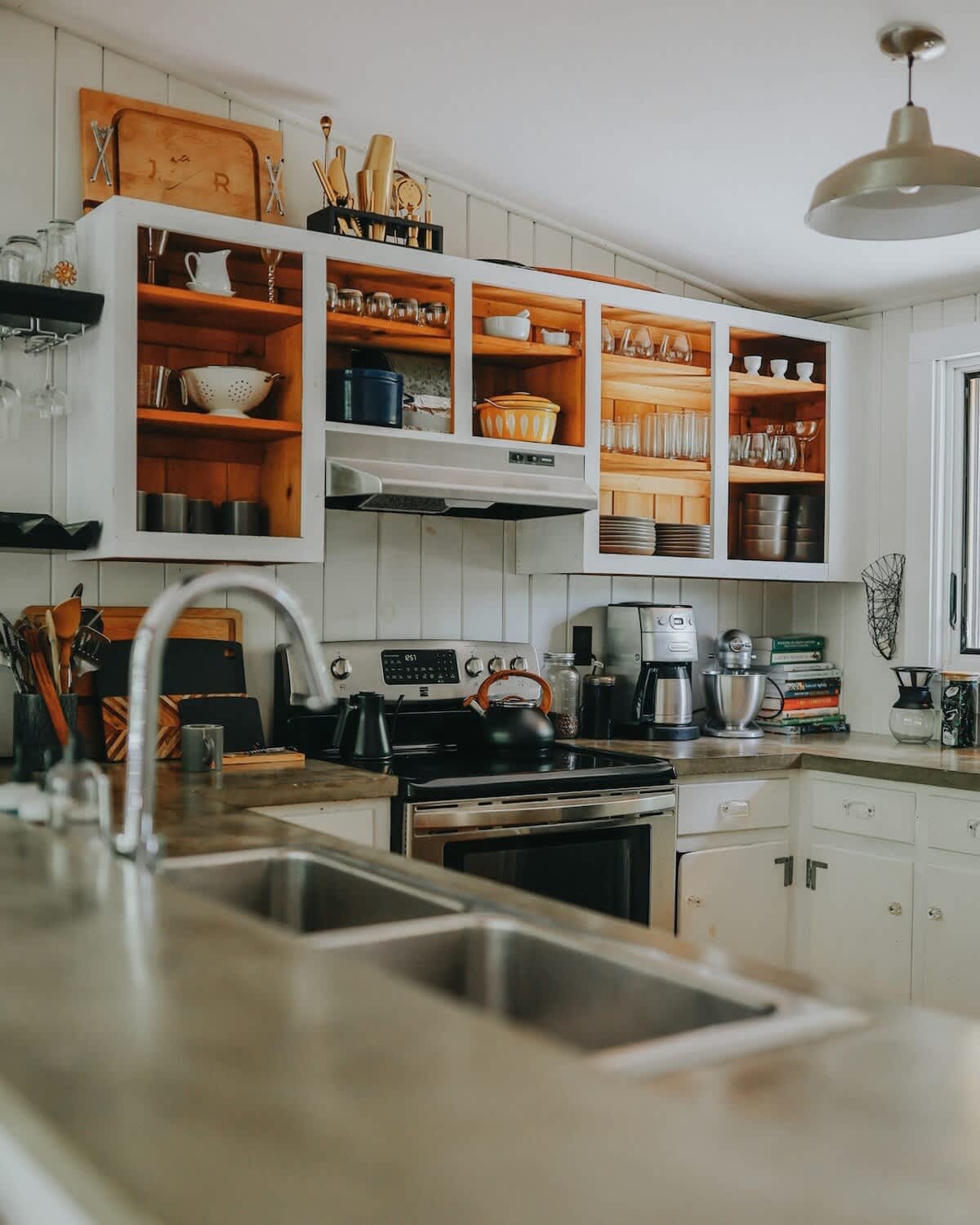 A rustic kitchen features white open shelving with natural wood interiors, displaying various dishes, glasses, and cooking utensils. A stainless steel sink and faucet are in the foreground, with a black stove and range hood visible in the background.