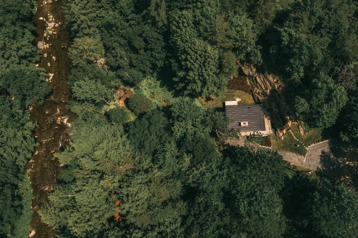 An aerial shot captures a dark-roofed cabin nestled within a vast, verdant forest, with a winding river flowing nearby. A stone path leads away from the cabin, and a small fire pit area is visible in a clearing.