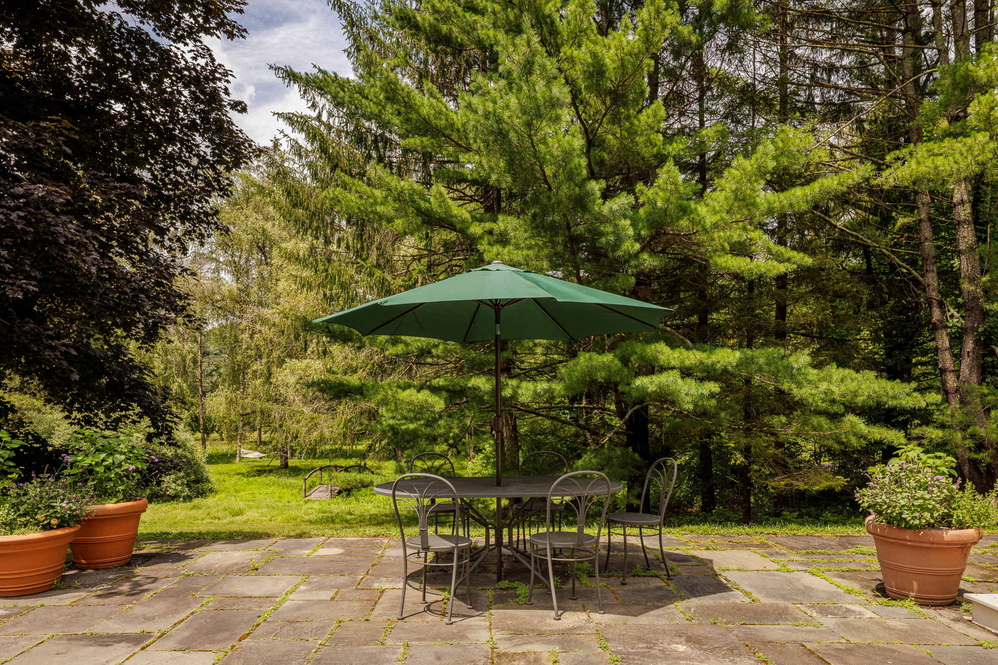 A stone patio shaded by a large green umbrella, featuring a metal outdoor dining table with four chairs. The patio is surrounded by lush greenery and tall trees, with potted plants adding decor.