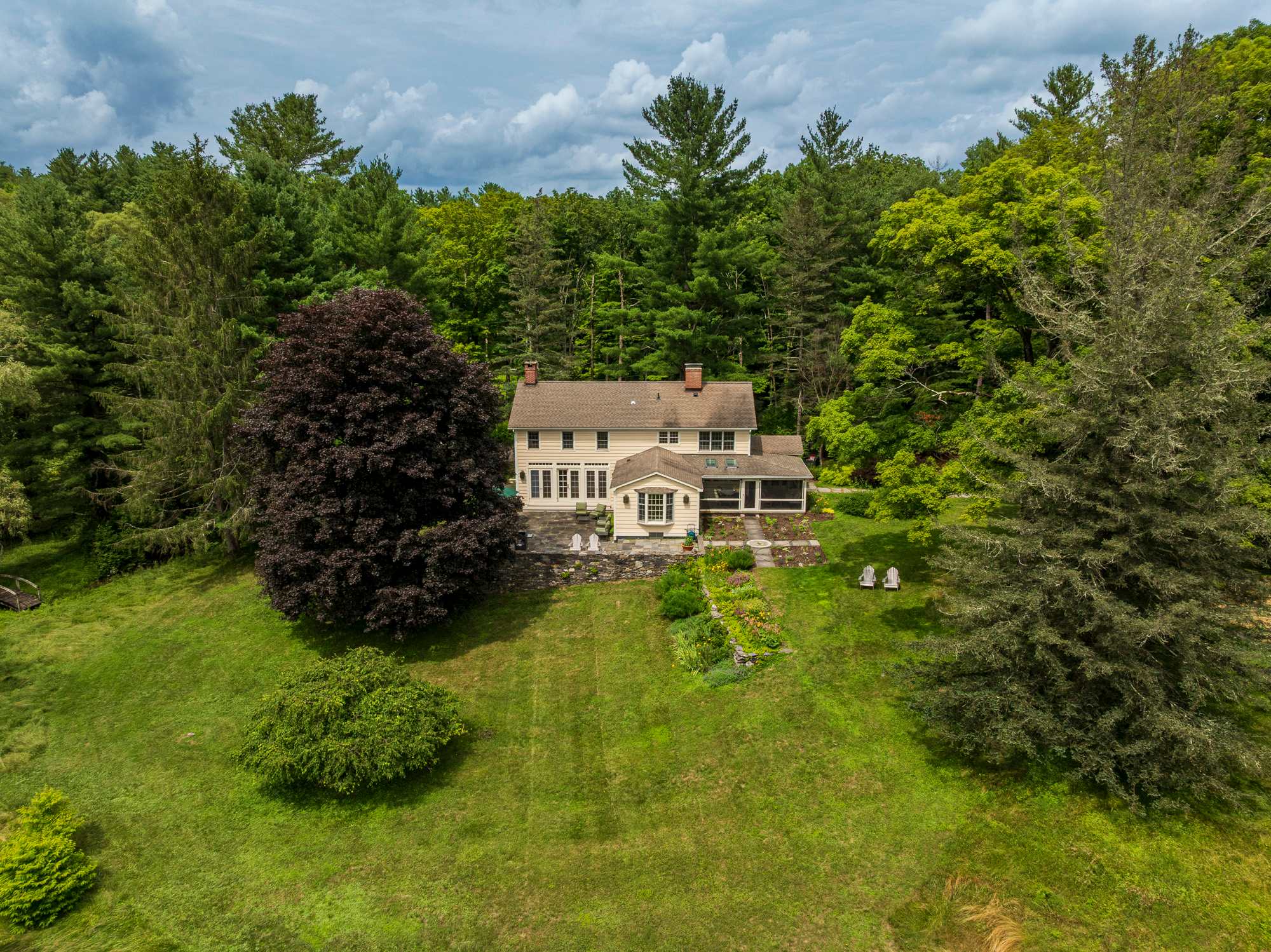 An aerial view showcases a light yellow house with dark shutters, nestled among dense green trees, featuring a large dark-leafed tree and a manicured lawn with outdoor seating.