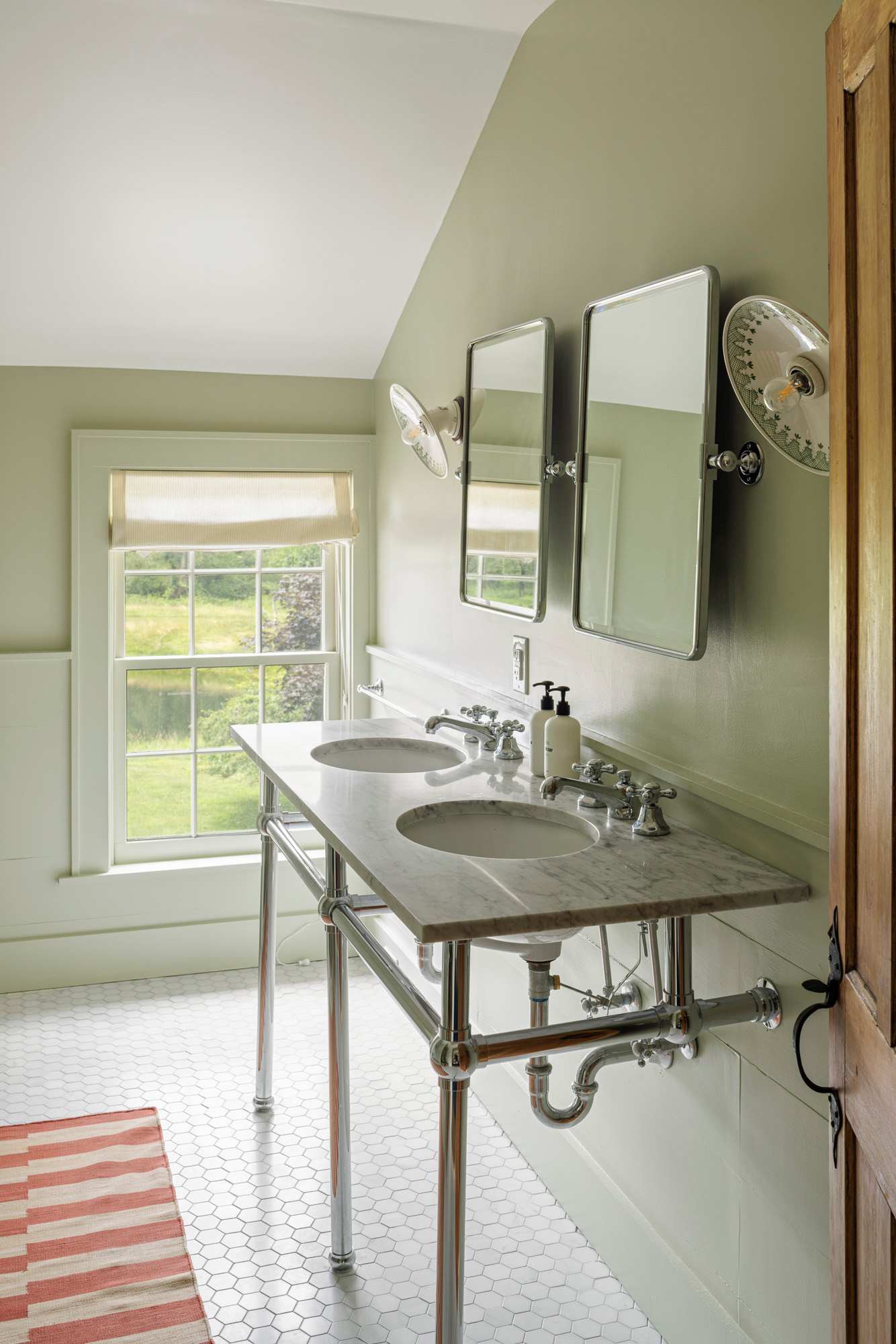A spacious bathroom with light green walls and white wainscoting, featuring a double vanity with a marble countertop and chrome legs. Two rectangular mirrors and wall-mounted lights are above the sinks, with a window offering an outdoor view.
