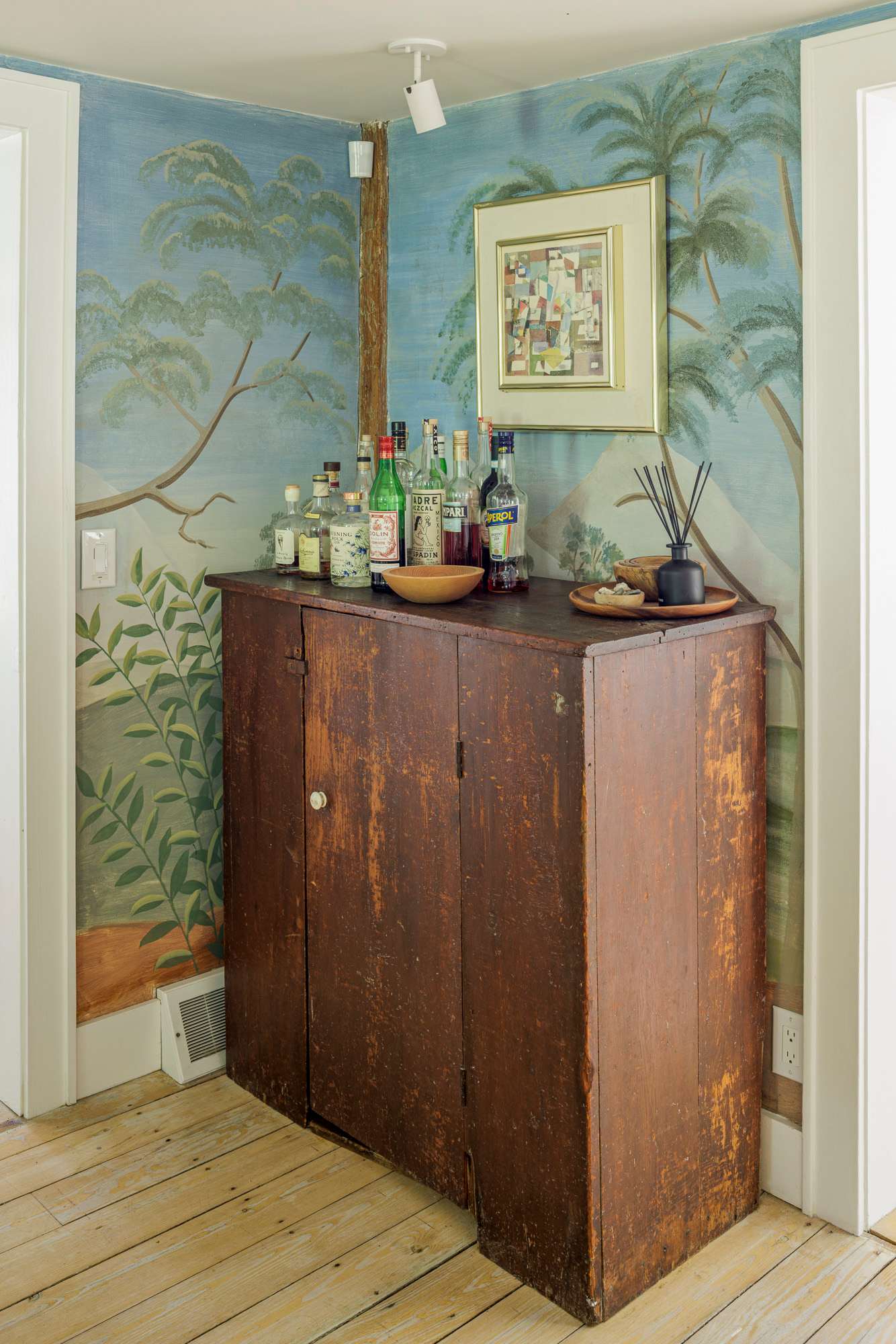 A rustic wooden cabinet serves as a bar area, topped with various liquor bottles, against a wall adorned with a scenic mural featuring trees and mountains. Light wood floors and a framed abstract artwork complete the eclectic space.