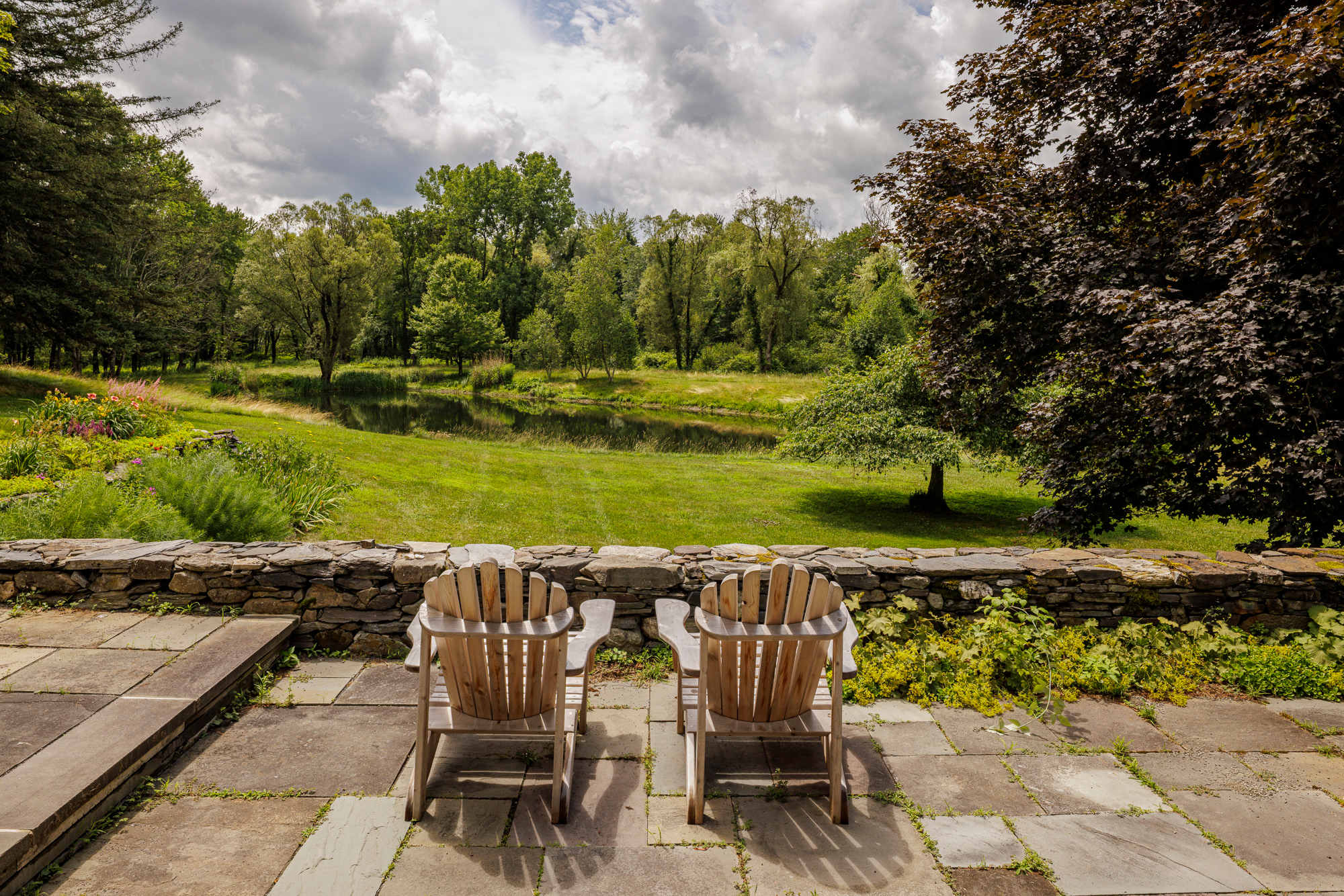 A stone patio with two wooden Adirondack chairs facing a picturesque view of a tranquil pond surrounded by lush green grass and mature trees under a cloudy sky. A low stone wall separates the patio from the landscape.