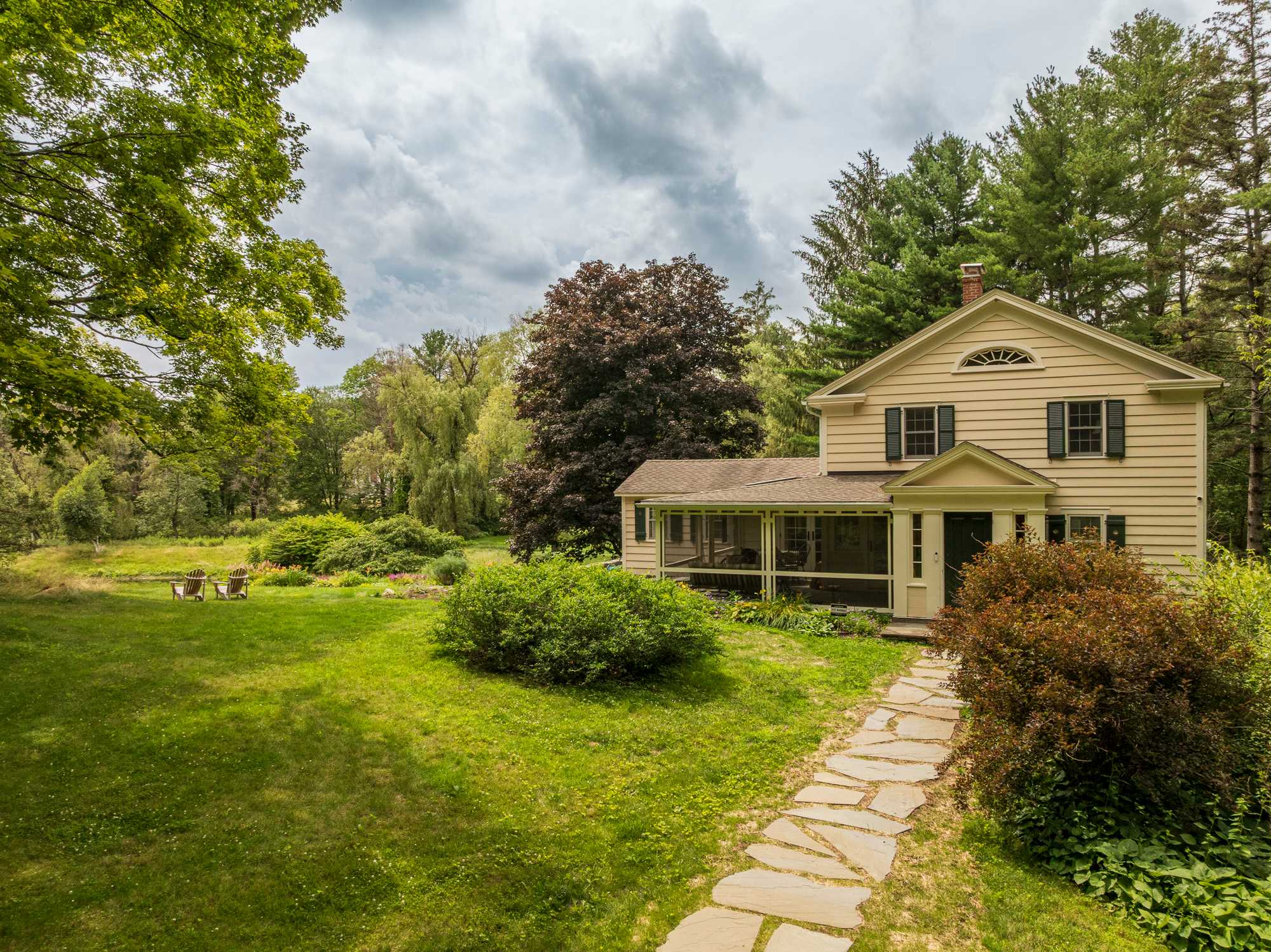 A light yellow house with dark shutters and a screened porch overlooks a lush green lawn and garden, with a stone path leading to outdoor seating.
