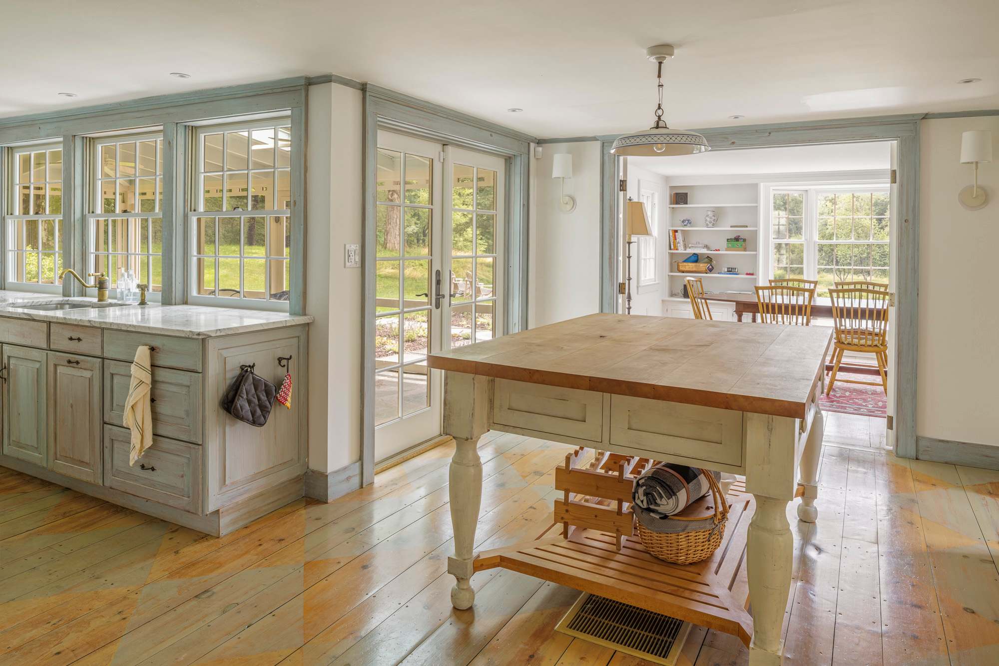 Bright kitchen with light blue-grey cabinets, marble countertops, and wide plank hardwood floors with a painted pattern. A large wooden island with storage sits in the center, leading to French doors and a dining area beyond.
