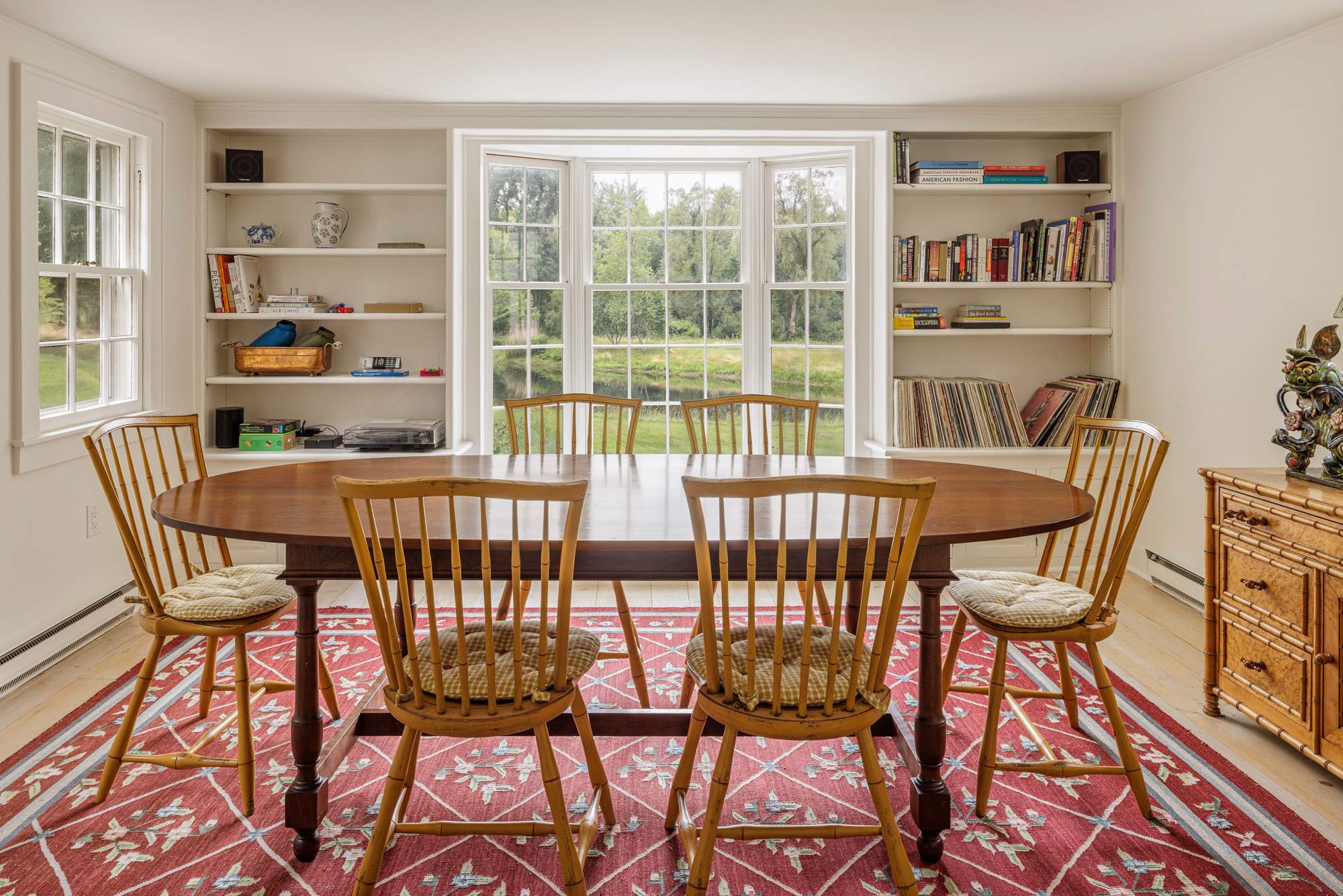 Elegant dining room with a large oval wooden table and six spindle-back chairs with patterned cushions, set on a red geometric rug. Built-in bookshelves flank a wide bay window offering views of the lush outdoor landscape.