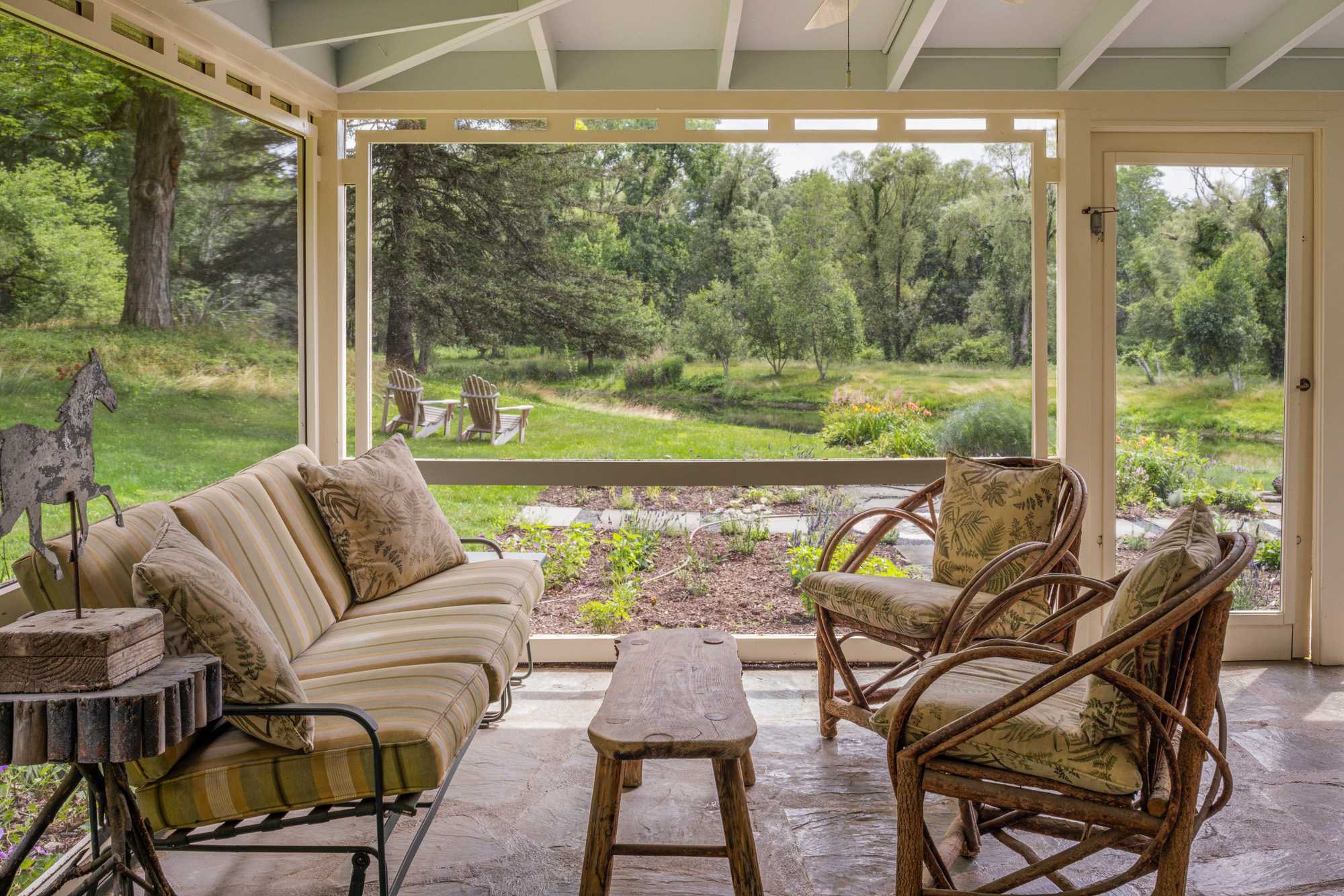 A spacious screened-in porch with flagstone flooring and comfortable wicker seating, offering an expansive view of the lush green yard and distant forest. The seating includes a sofa and two armchairs with patterned cushions.