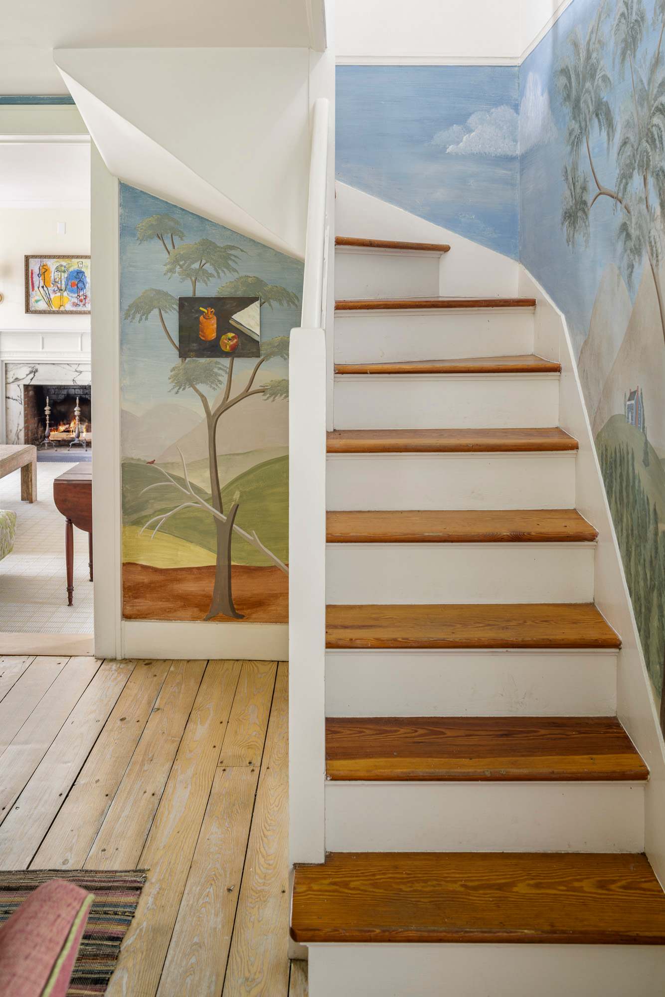 A charming entryway featuring a wooden staircase with white risers and natural wood treads, set against walls adorned with a whimsical hand-painted mural depicting trees and a landscape. A glimpse of the living room fireplace is visible beyond.