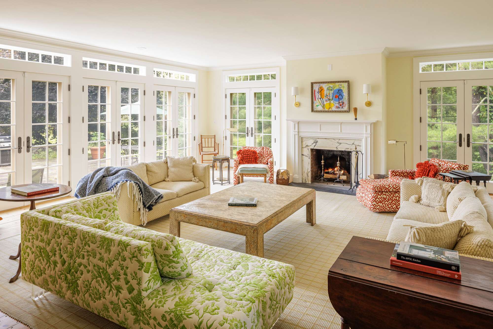 Spacious living room featuring multiple French doors that open to the outdoors, a classic white fireplace, and a mix of patterned sofas and armchairs. A large wooden coffee table anchors the room, which is covered with a light patterned rug.