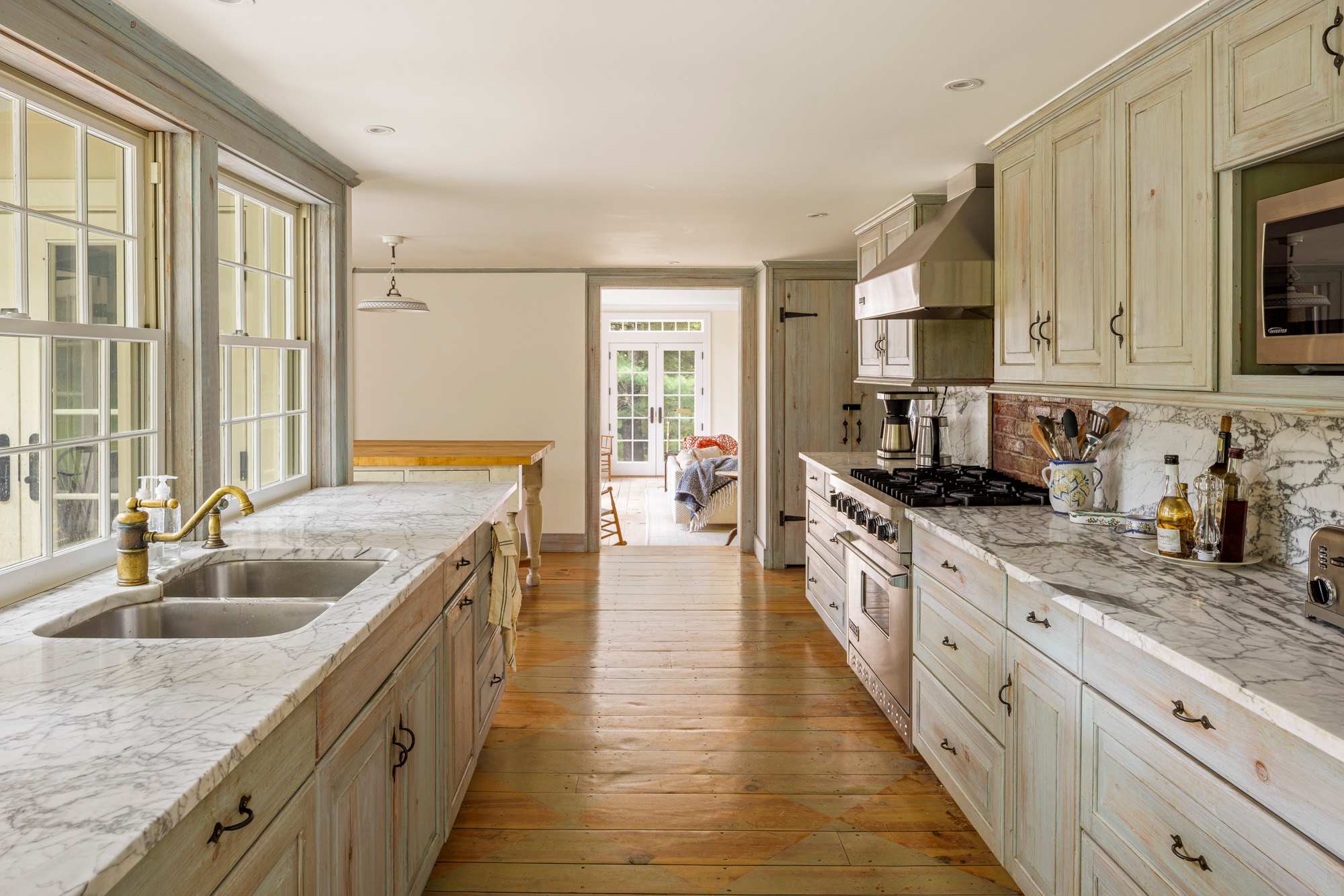 Long galley kitchen featuring light blue-grey distressed wood cabinets, white marble countertops, and wide plank hardwood floors. A stainless steel range and hood are visible, with a double sink under a window overlooking the outdoors.