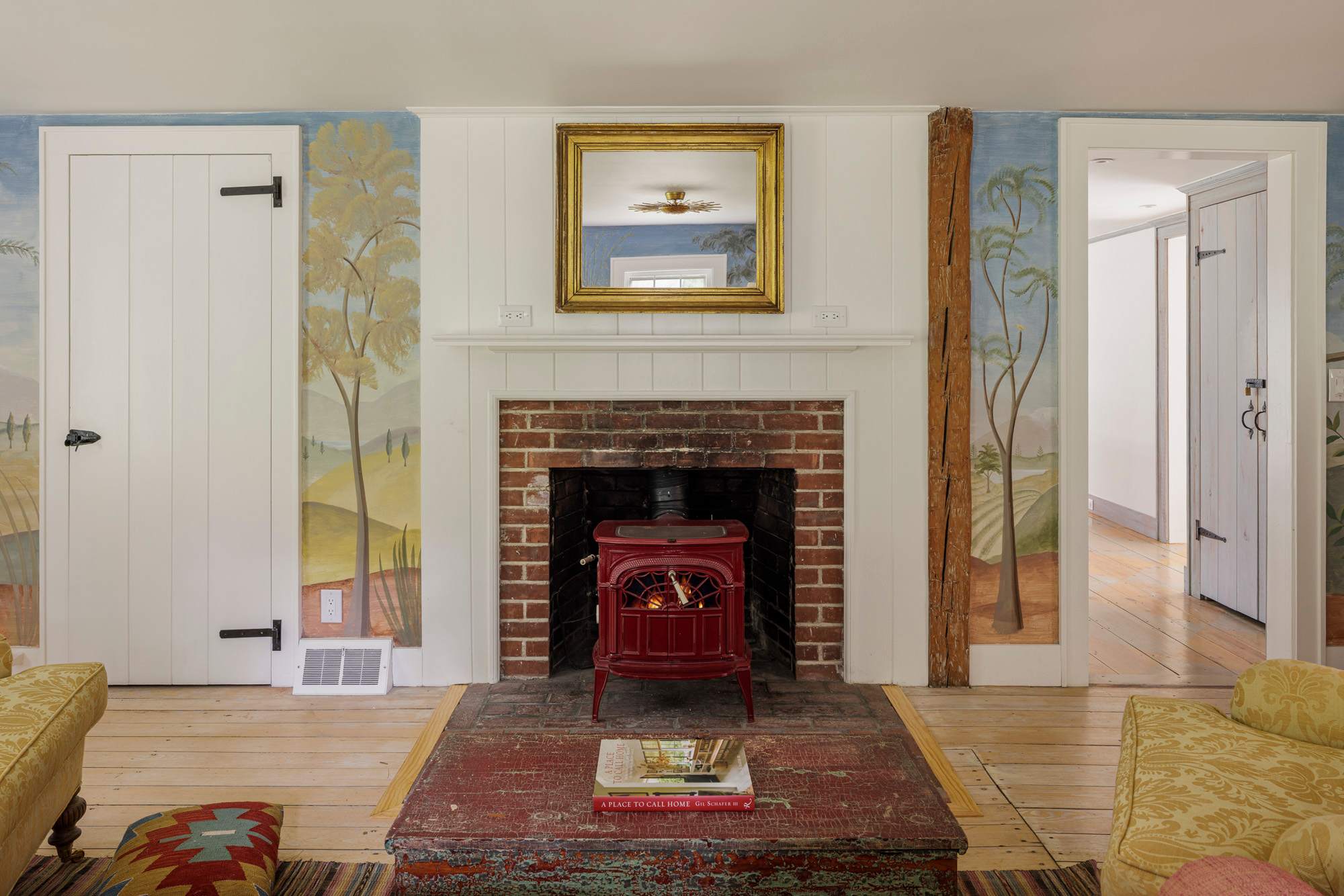 A cozy living space with hand-painted mural walls depicting a pastoral landscape, centered around a brick fireplace with a red wood-burning stove. Two patterned sofas flank a rustic coffee table, and a golden mirror hangs above the mantel.