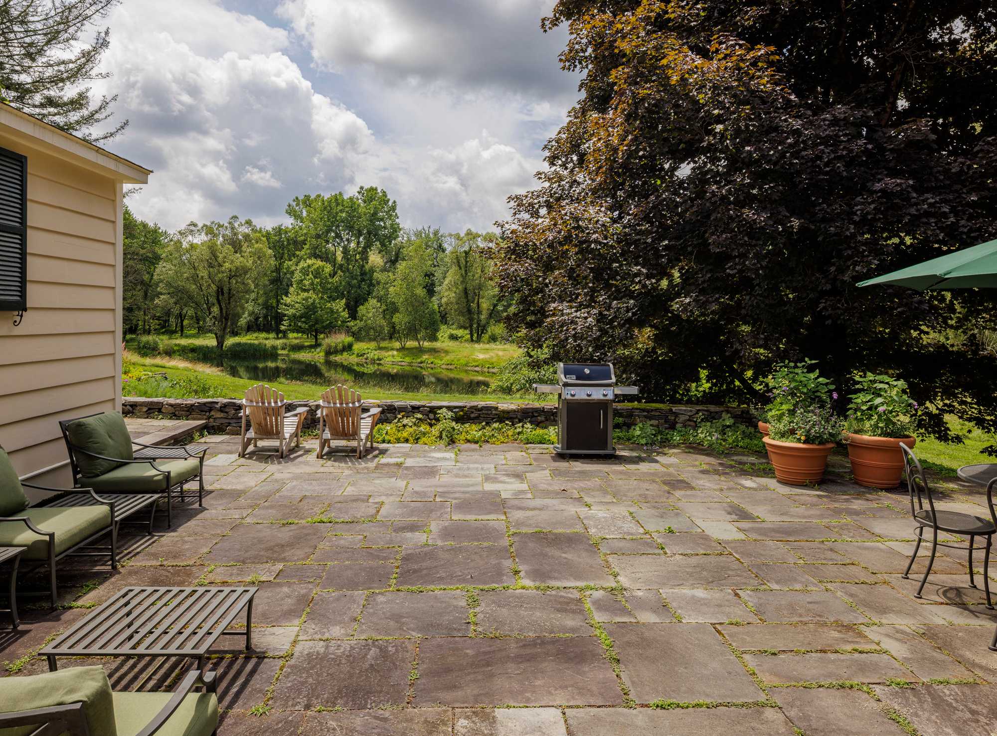 A spacious stone patio with a BBQ grill, two wooden Adirondack chairs, and a metal outdoor seating area with green cushions. The patio overlooks a lush green landscape with a pond and trees.
