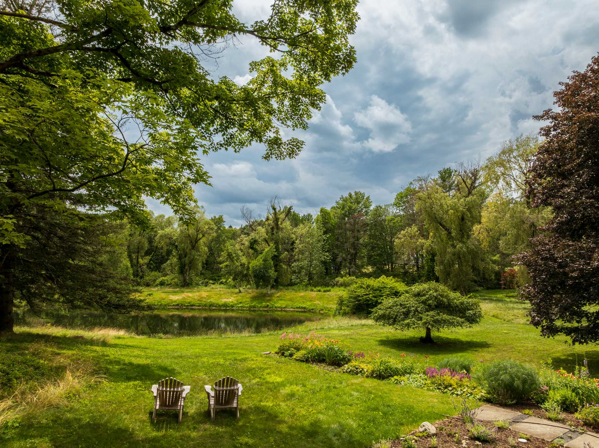 A picturesque view of a lush green yard with two Adirondack chairs facing a tranquil pond and dense forest, framed by mature trees overhead. Colorful flower beds add a touch of vibrancy to the natural landscape.