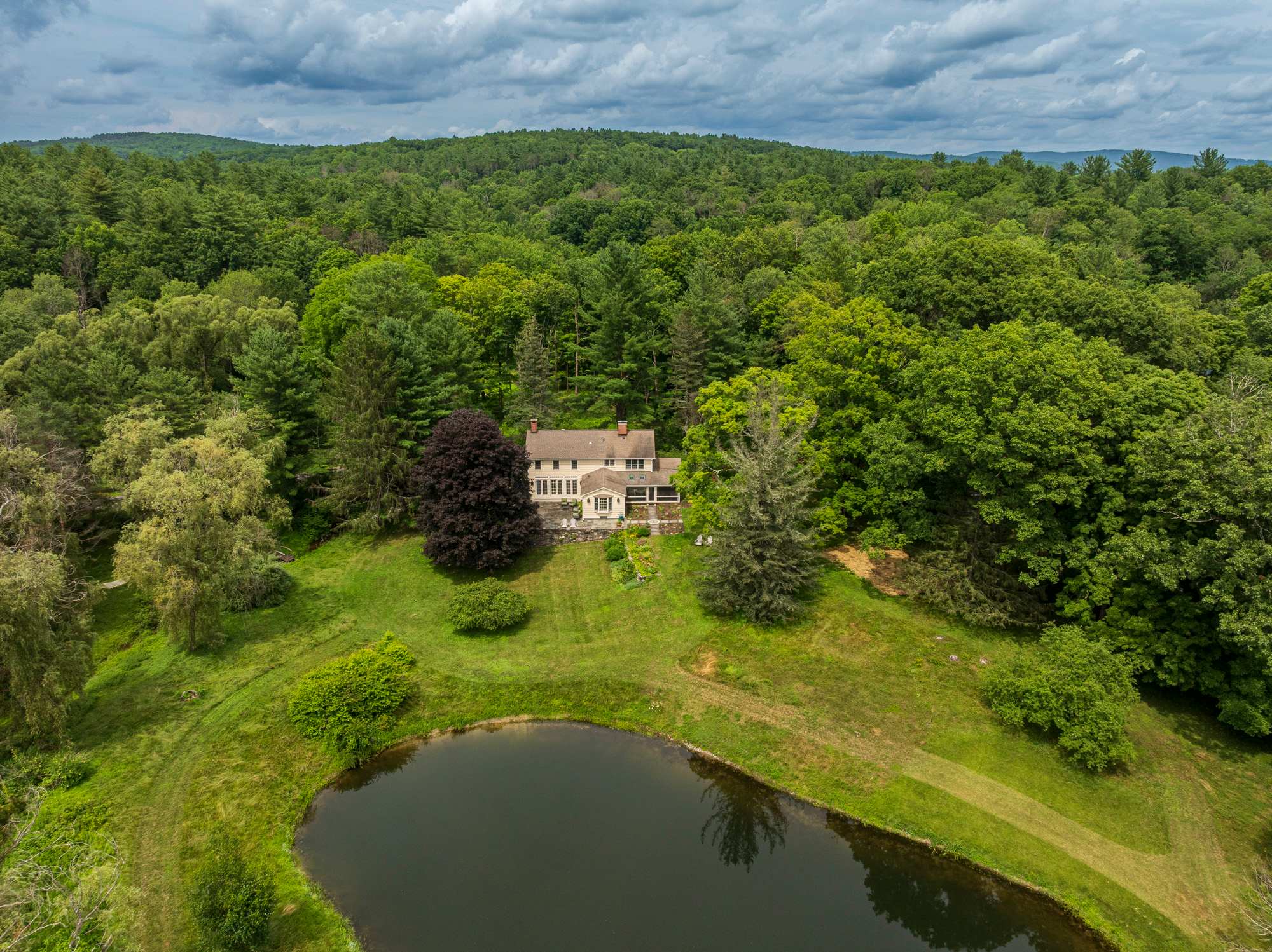 An aerial view showcases the light yellow house nestled amidst a vast expanse of green forest and rolling hills, with a large, dark pond in the foreground reflecting the cloudy sky. The property offers extensive natural beauty and privacy.