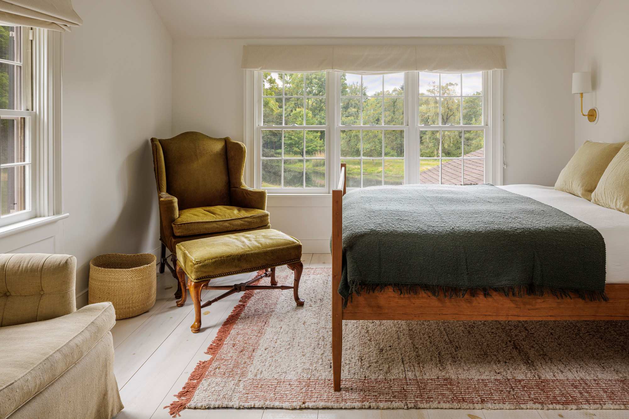 A bright bedroom with white walls, light wood floors, and a large window overlooking a lush green landscape. Features a wooden bed frame with a dark green blanket, a vintage green armchair with an ottoman, and a woven basket.