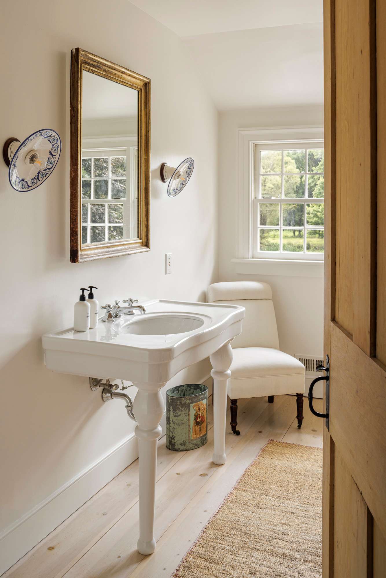 A bright bathroom with white walls and light wood floors, featuring a classic white pedestal sink with chrome fixtures and a gold-framed mirror. Decorative wall sconces flank the mirror, and a window provides natural light and a view of the outdoors.