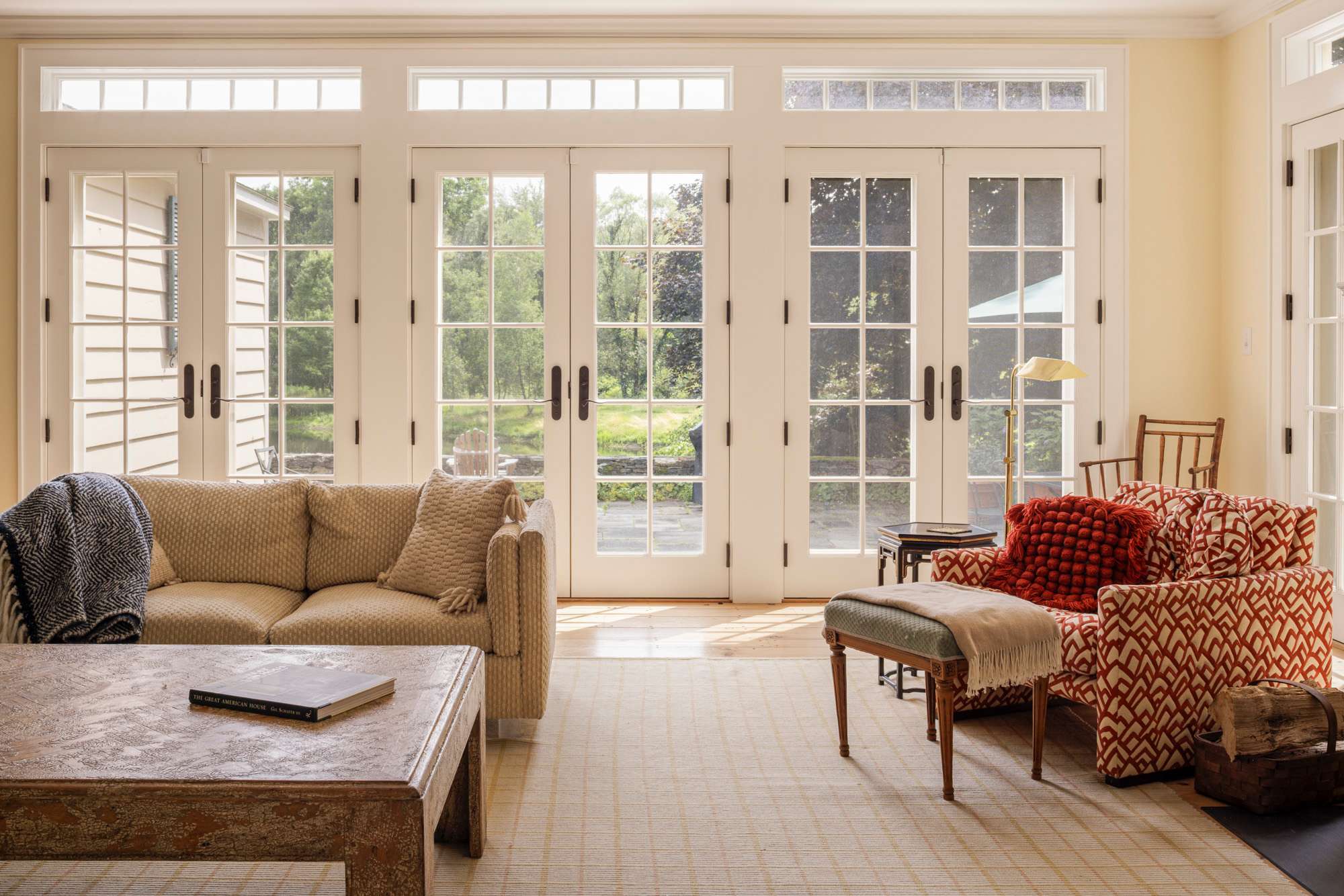 Bright living room with several sets of French doors and transom windows allowing abundant natural light and views of the garden. Two comfortable sofas and an armchair with a patterned throw are arranged around a large wooden coffee table.
