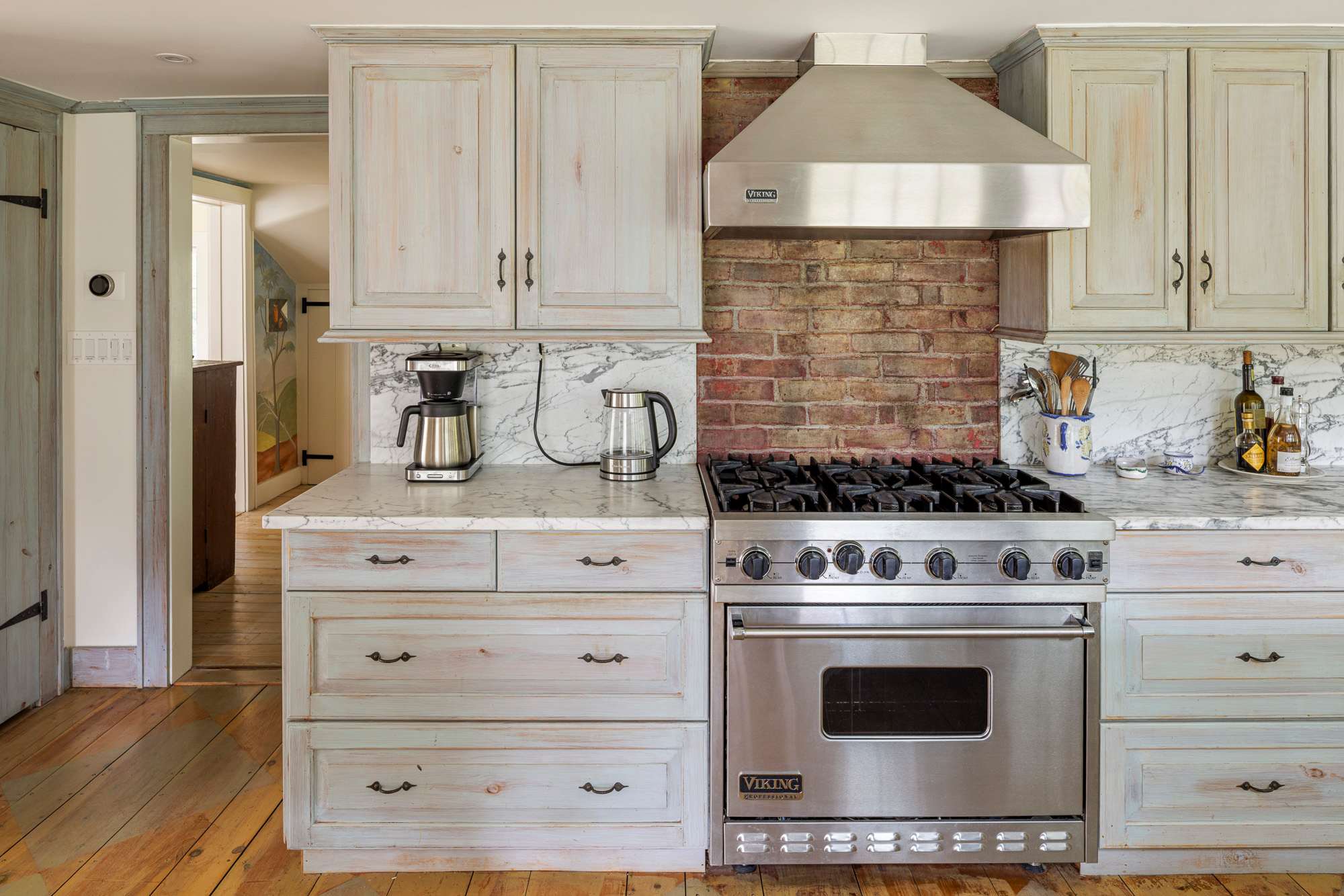 Rustic kitchen with light blue-grey distressed wood cabinets, white marble countertops, a stainless steel Viking range, and an exposed brick backsplash under a stainless steel range hood. Hardwood floors complete the warm, inviting look.