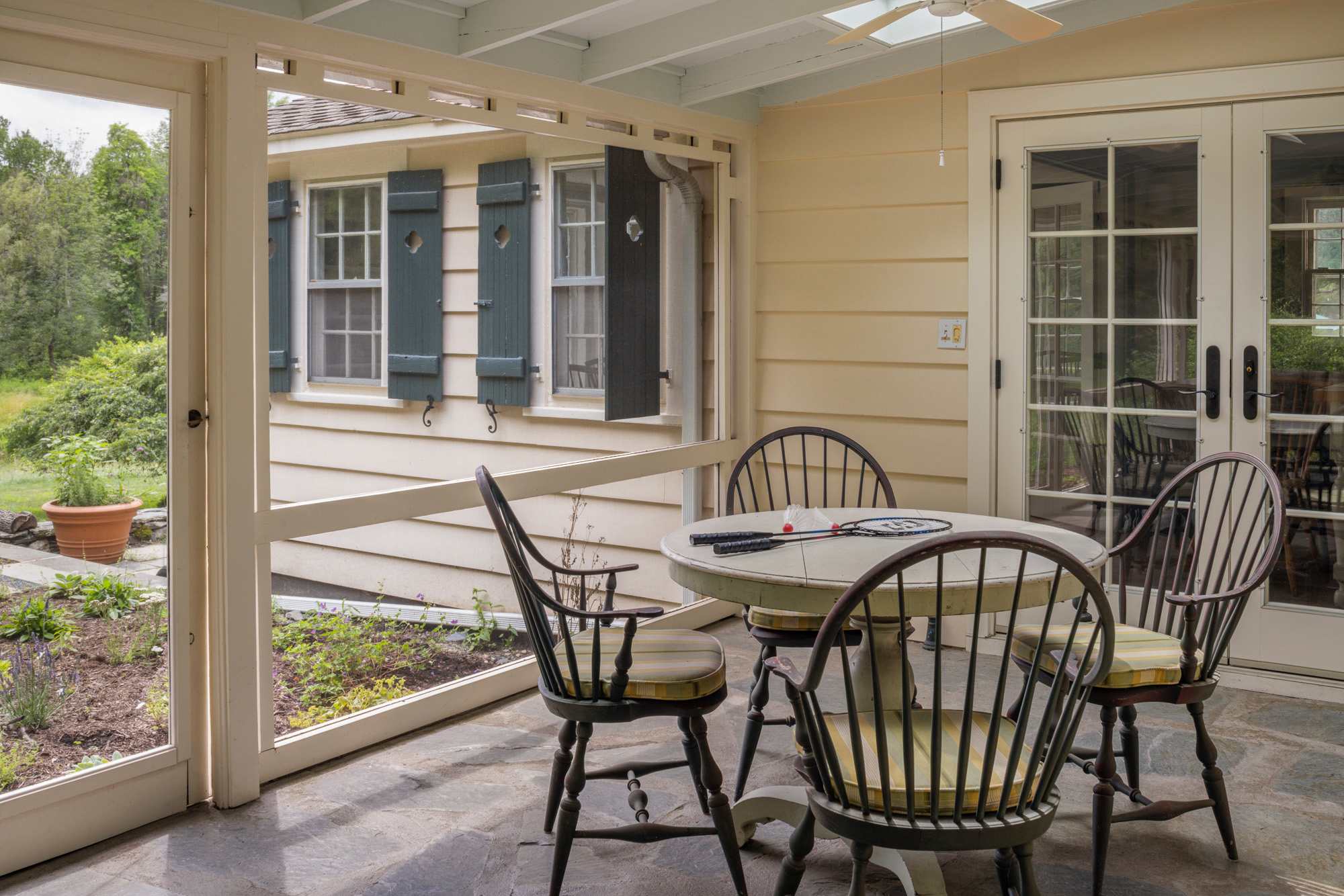 A screened-in porch with a round dining table and four wooden chairs with striped cushions, set on a stone tile floor. The porch overlooks a garden area and a house with blue shutters.