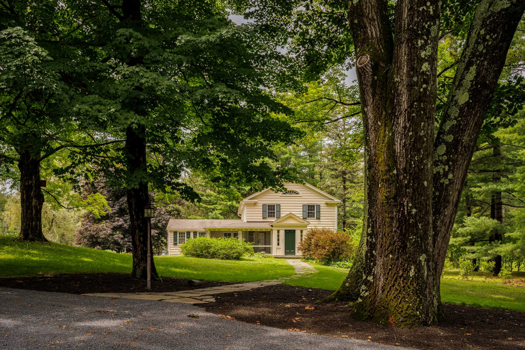 The front of a light yellow house with dark green shutters and a matching front door is framed by large, mature trees and a stone pathway leading to a screened porch.